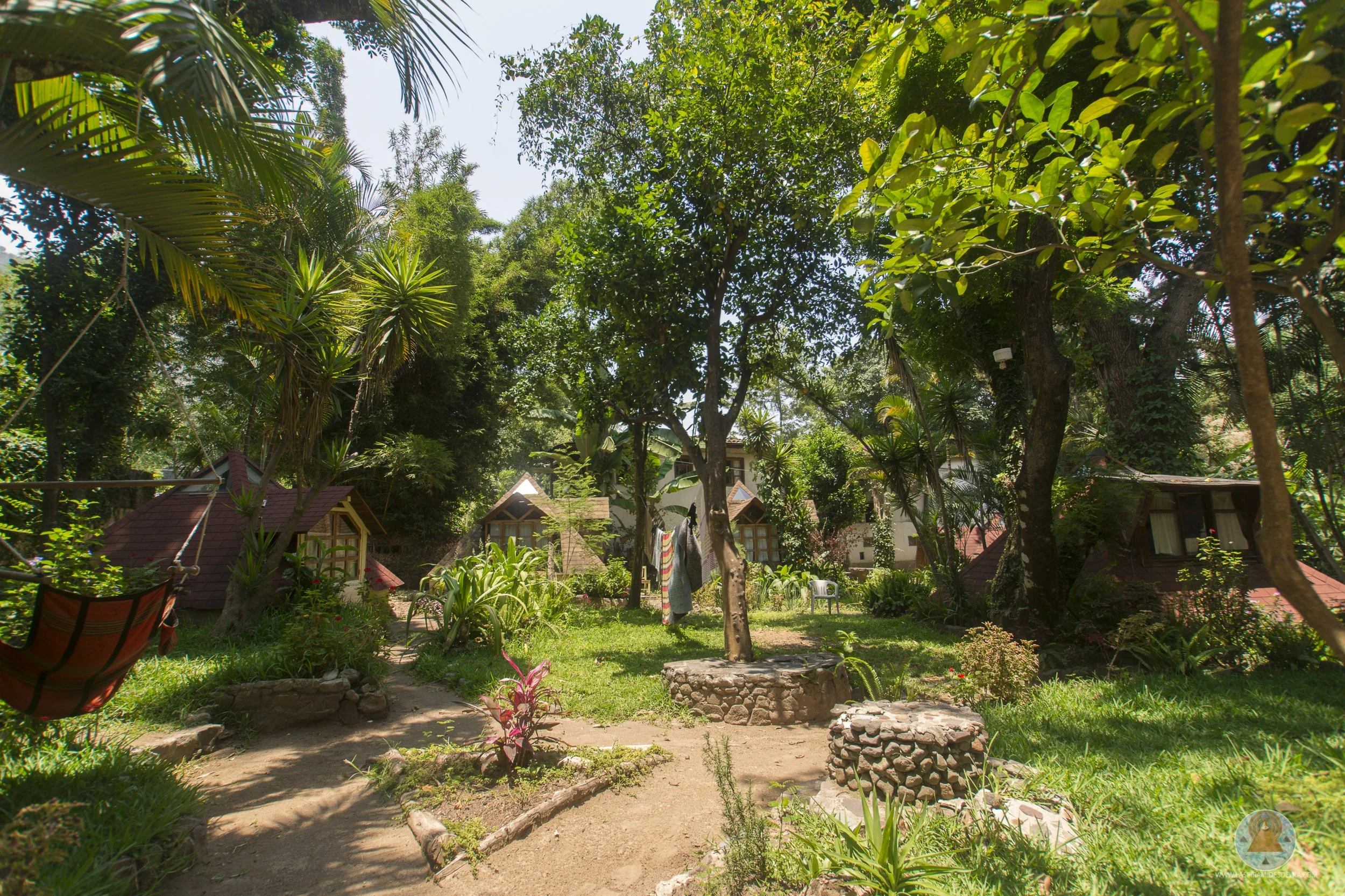 Jardín con árboles grandes, plantas tropicales, casas pequeñas con techos rojos, y muebles de jardín, en un día soleado