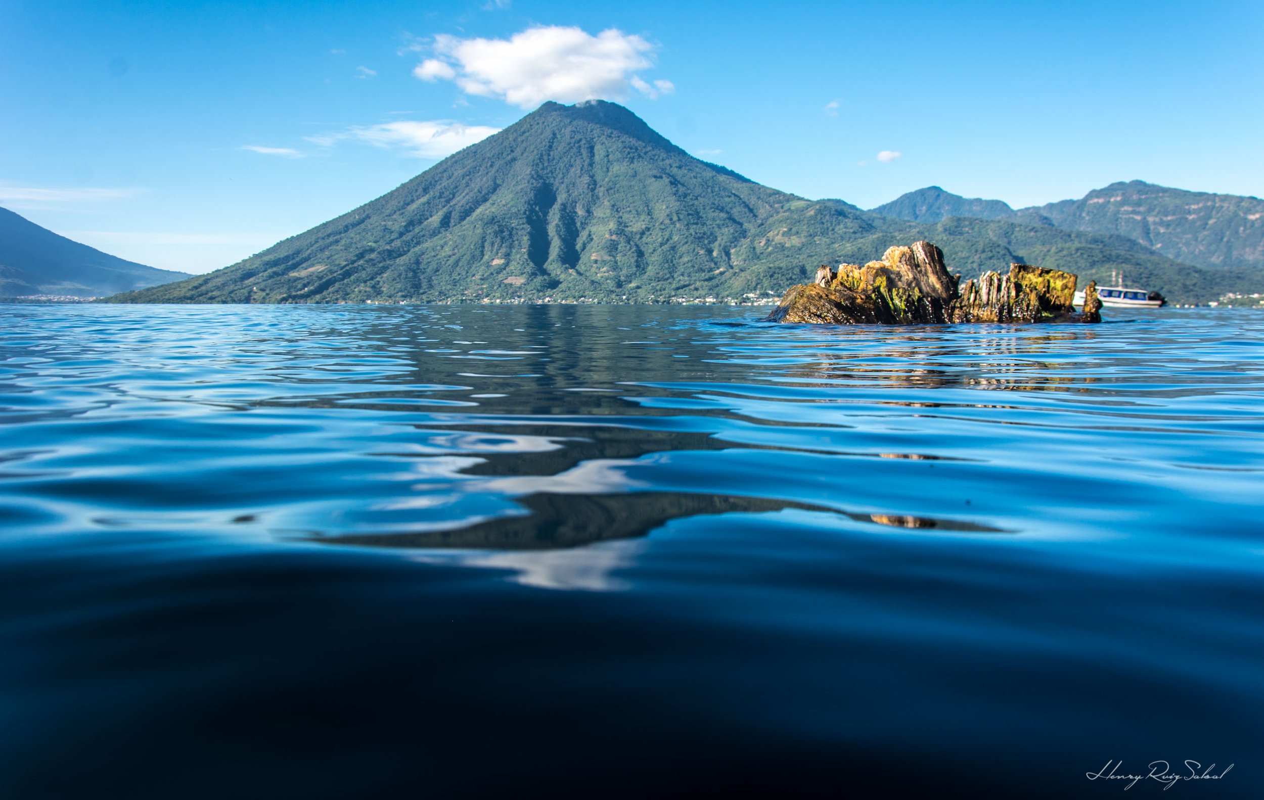 Un lago con una isla rocosa y un volcán grande en el fondo, rodeado de montañas y un cielo azul con algunas nubes.