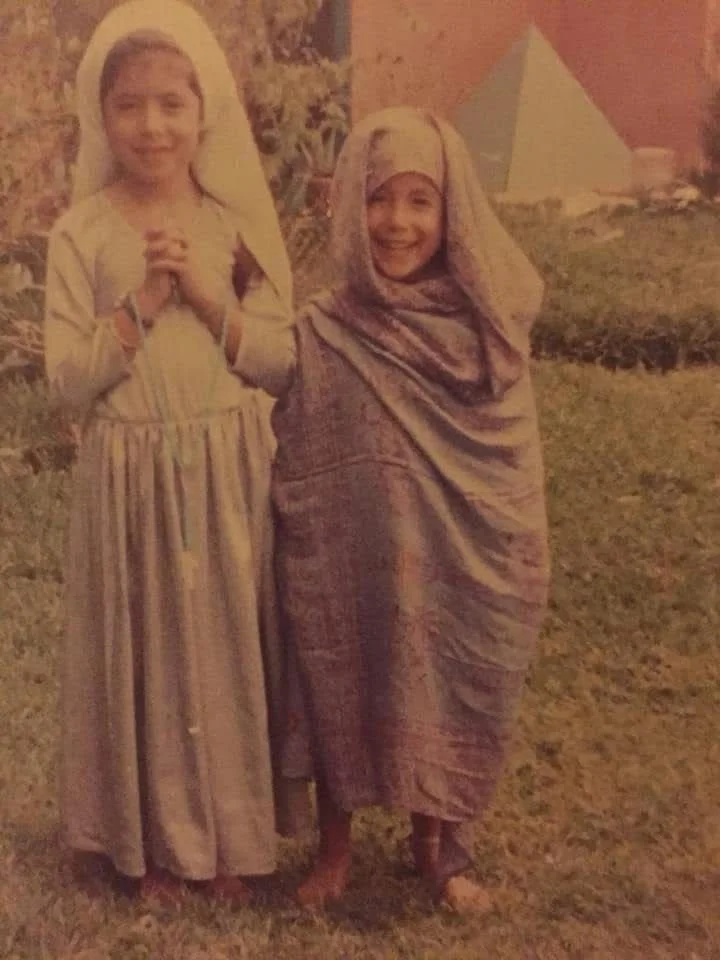 Dos niñas vestidas con ropas tradicionales de origen africano, una con ropas de color claro y la otra con ropa de tonos lavanda, están de pie en un jardín, ambas sonrisas y parecen felices.