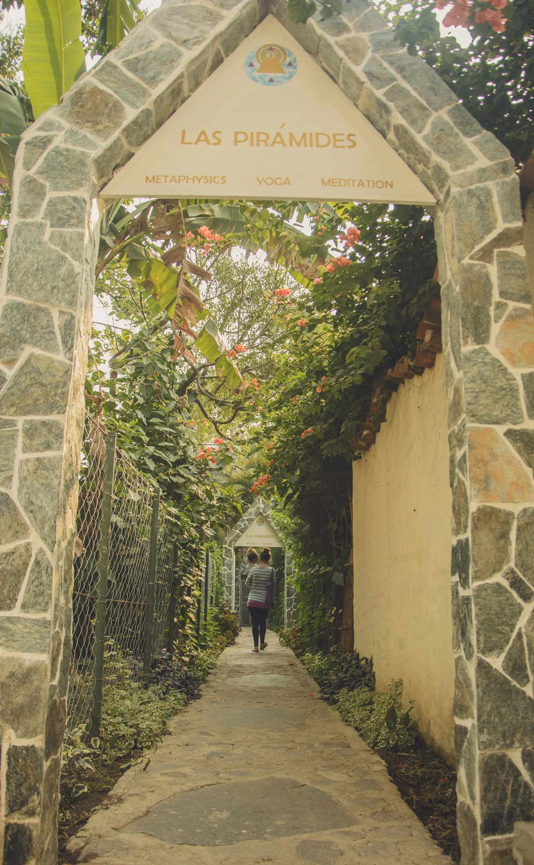 Entrada a un sendero rodeado de vegetación, con arcos de piedra y carteles que dicen 'Las Pirámides', ofreciendo actividades como metafísica, yoga y meditación, con dos personas caminando por el sendero.
