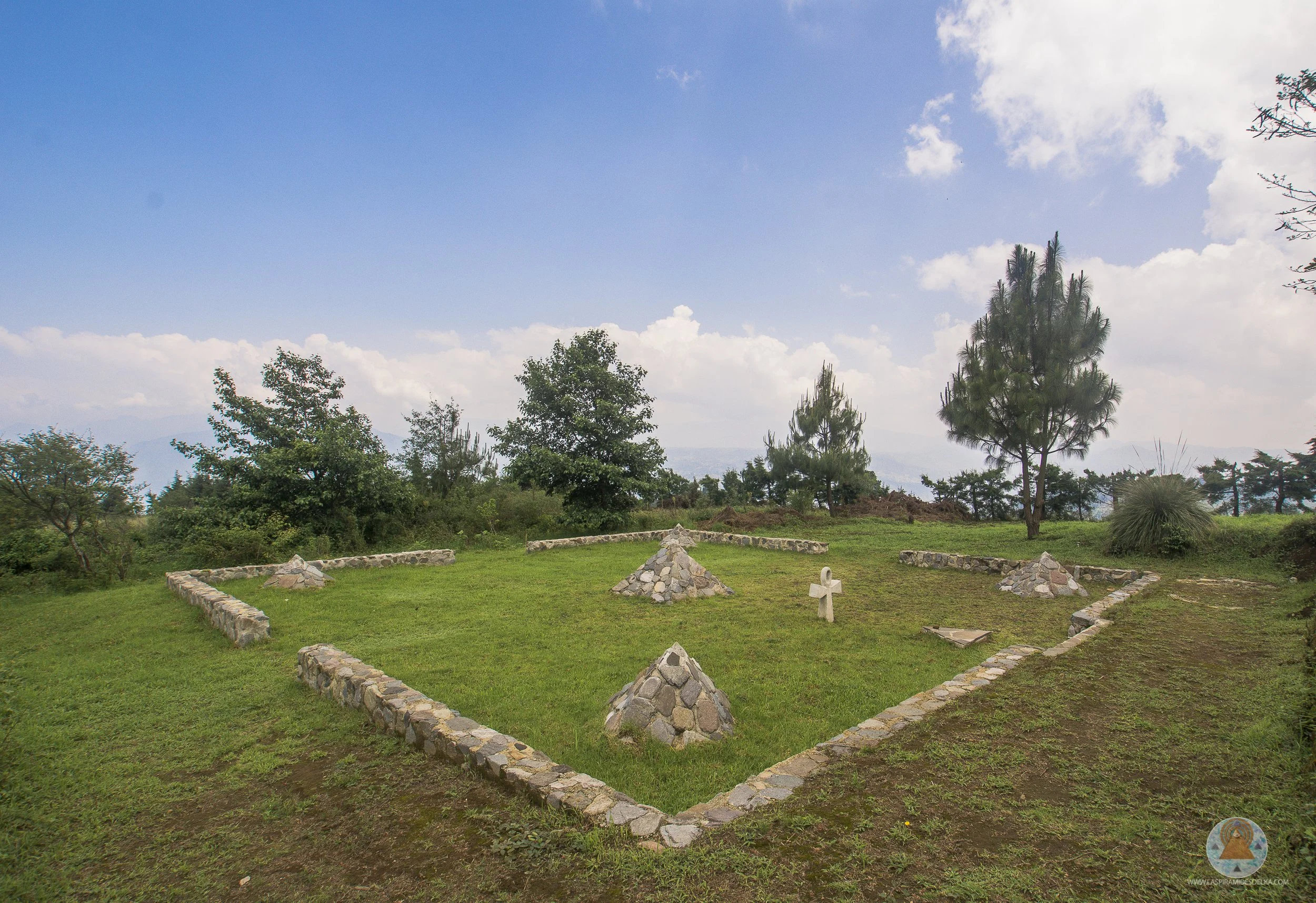 Vista de un jardín de cementerio con túmulos de piedra, rodeado de árboles y un cielo despejado con algunas nubes.