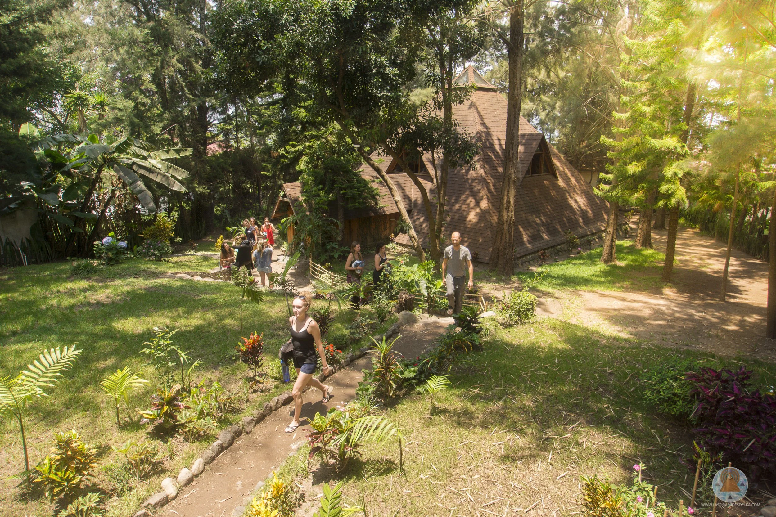 Grupo de personas caminando por un sendero en un entorno natural con árboles y una casa de estilo rústico de madera en el fondo.