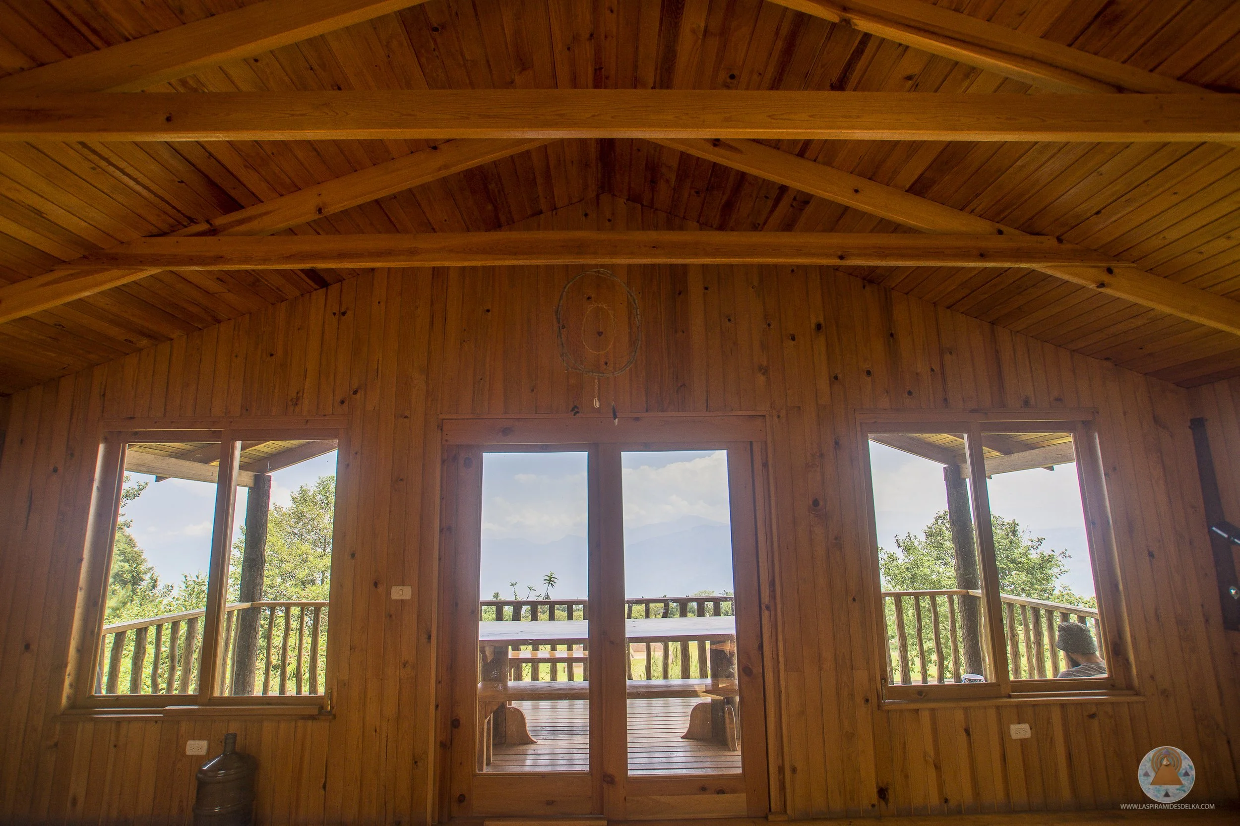 Interiores de una cabaña de madera con ventanas grandes que ofrecen vista a la naturaleza y un cielo despejado.