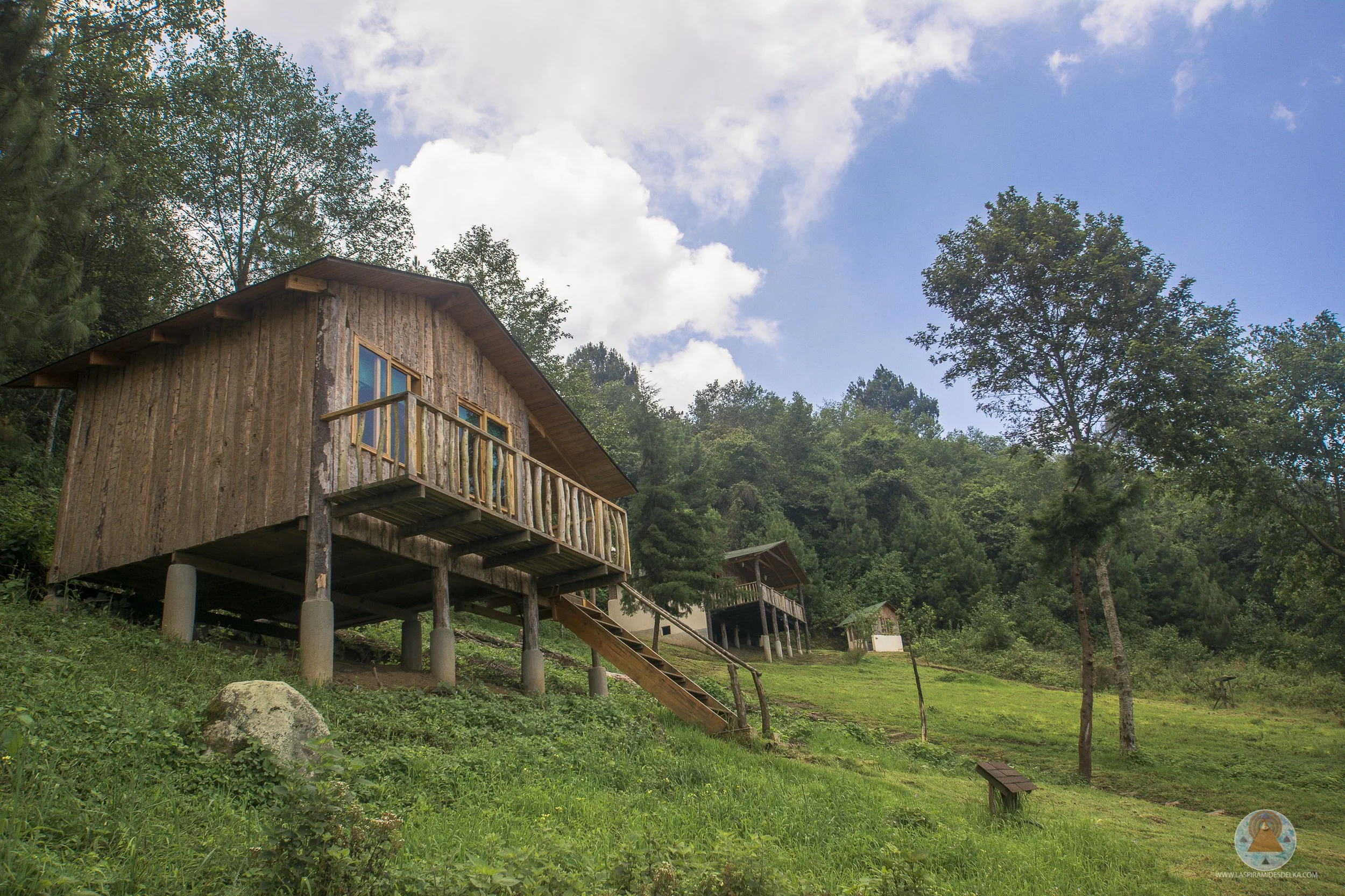 Casa de madera elevada en columnas en una zona boscosa, con árboles altos y cielo parcialmente nublado.