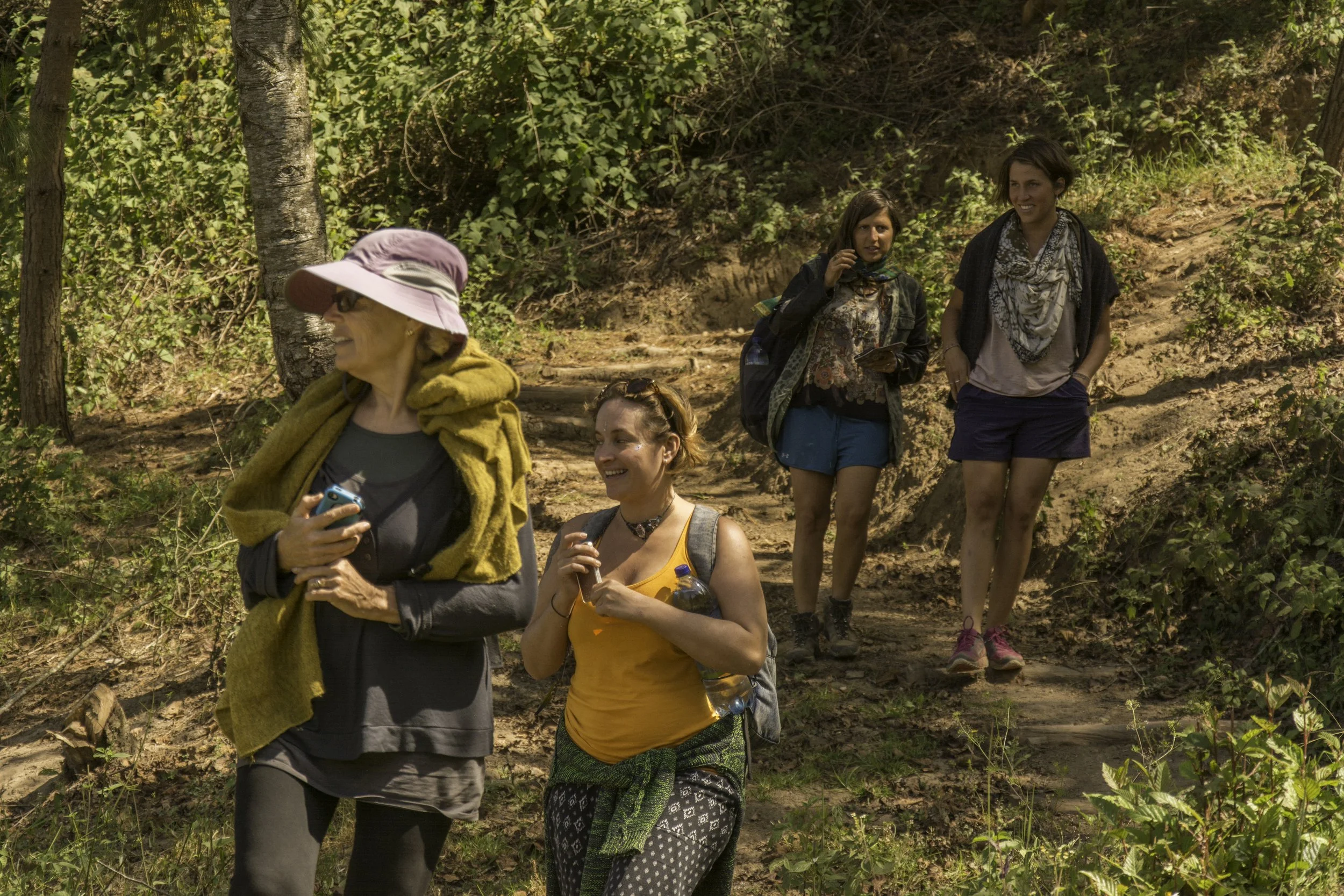 Grupo de personas caminando por un sendero en un bosque, disfrutando de un día soleado. Algunas llevan mochilas y ropa deportiva, parecen estar en una excursión o caminata.