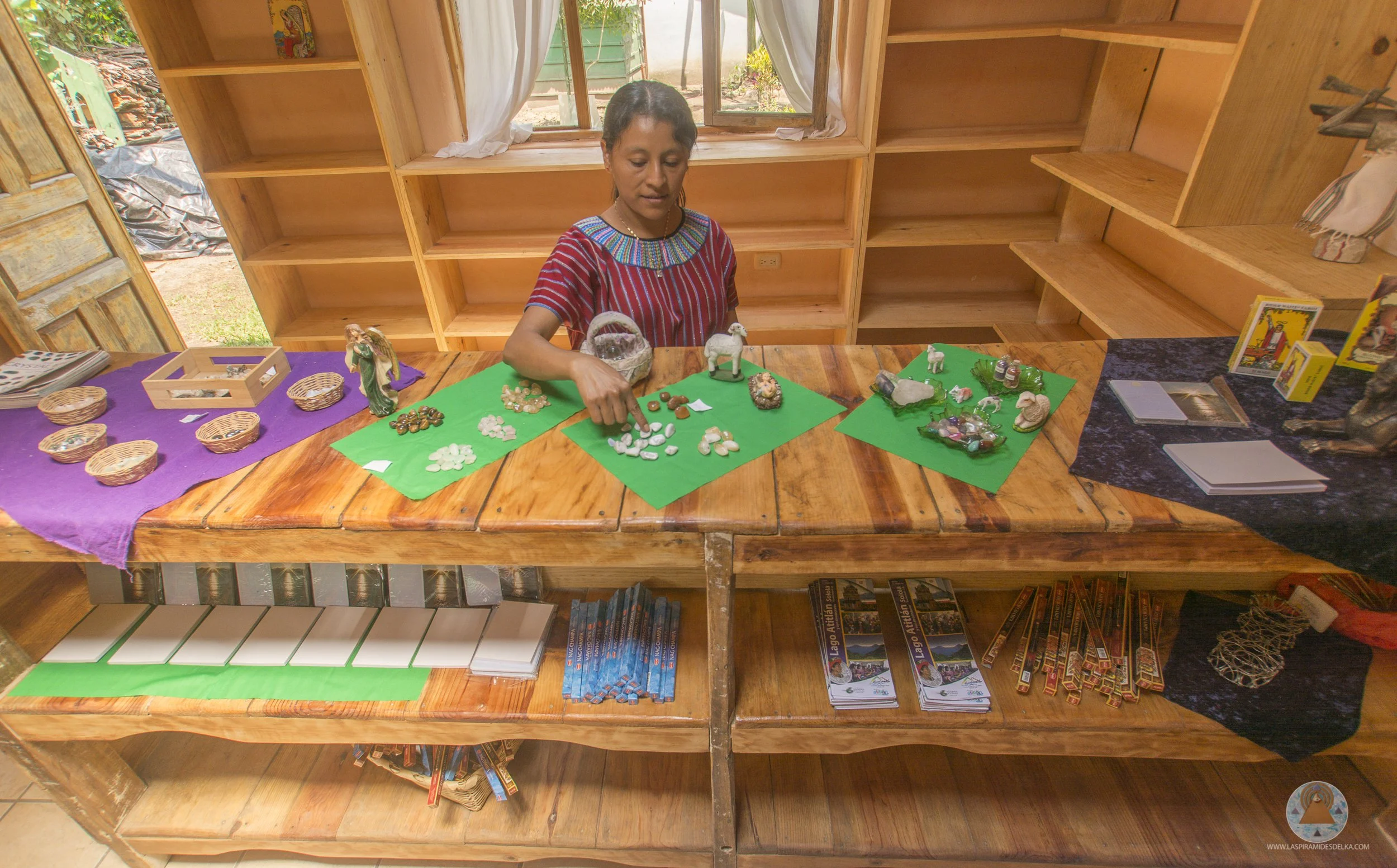 Mujer indígena mexicana en una habitación de madera, exhibiendo piedras y minerales en mesas cubiertas con papel verde y morado.