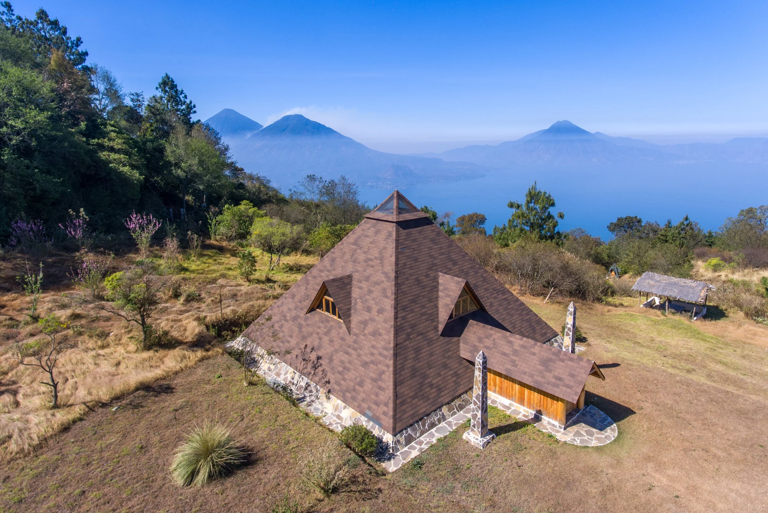 Casa en un paisaje montañoso con volcán y lago en el fondo, rodeada de árboles y vegetación.
