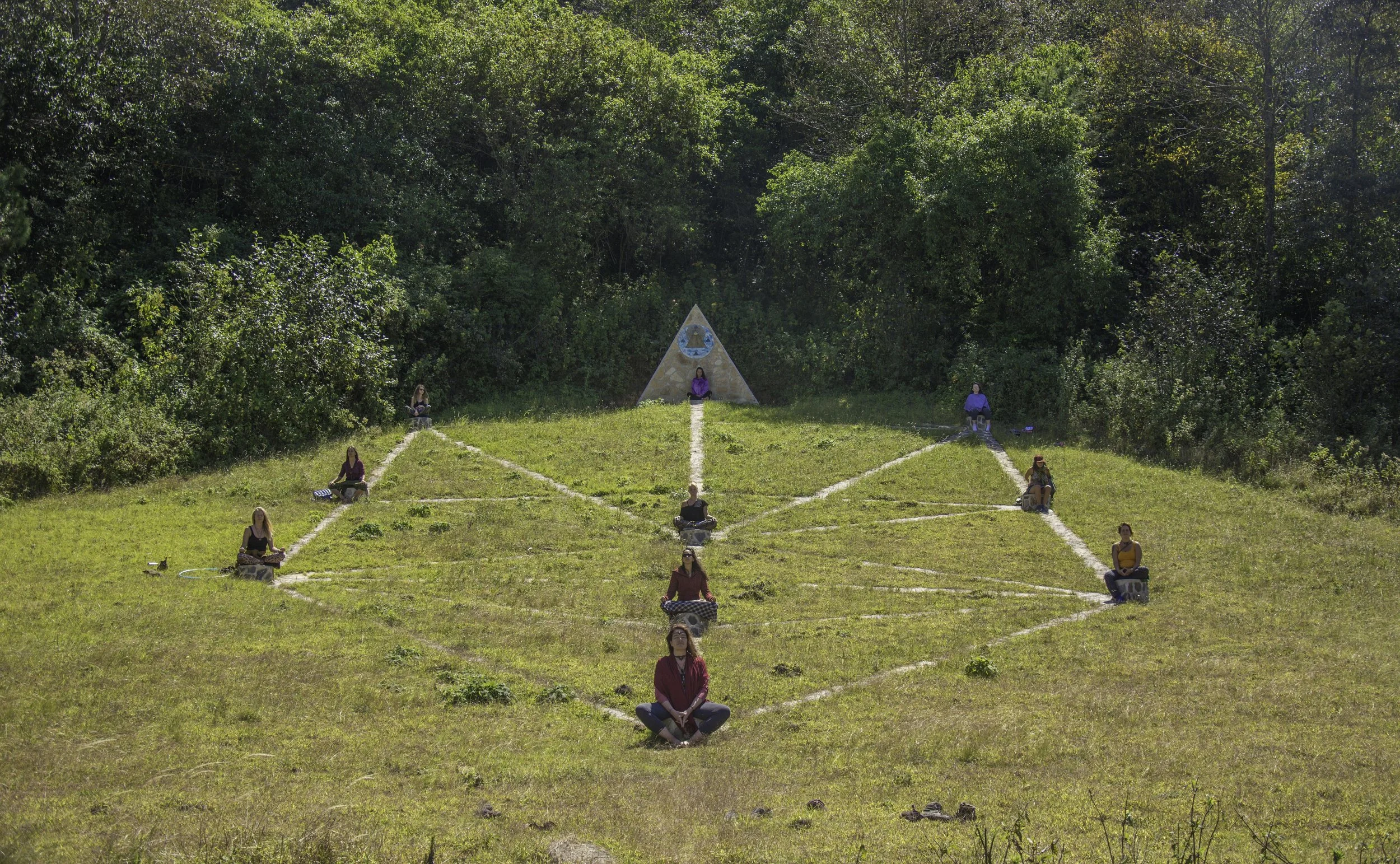 Grupo de personas meditando en un campo, sentados en posición de loto con distanciamiento, con un triángulo de piedra en el fondo y rodeados de vegetación.