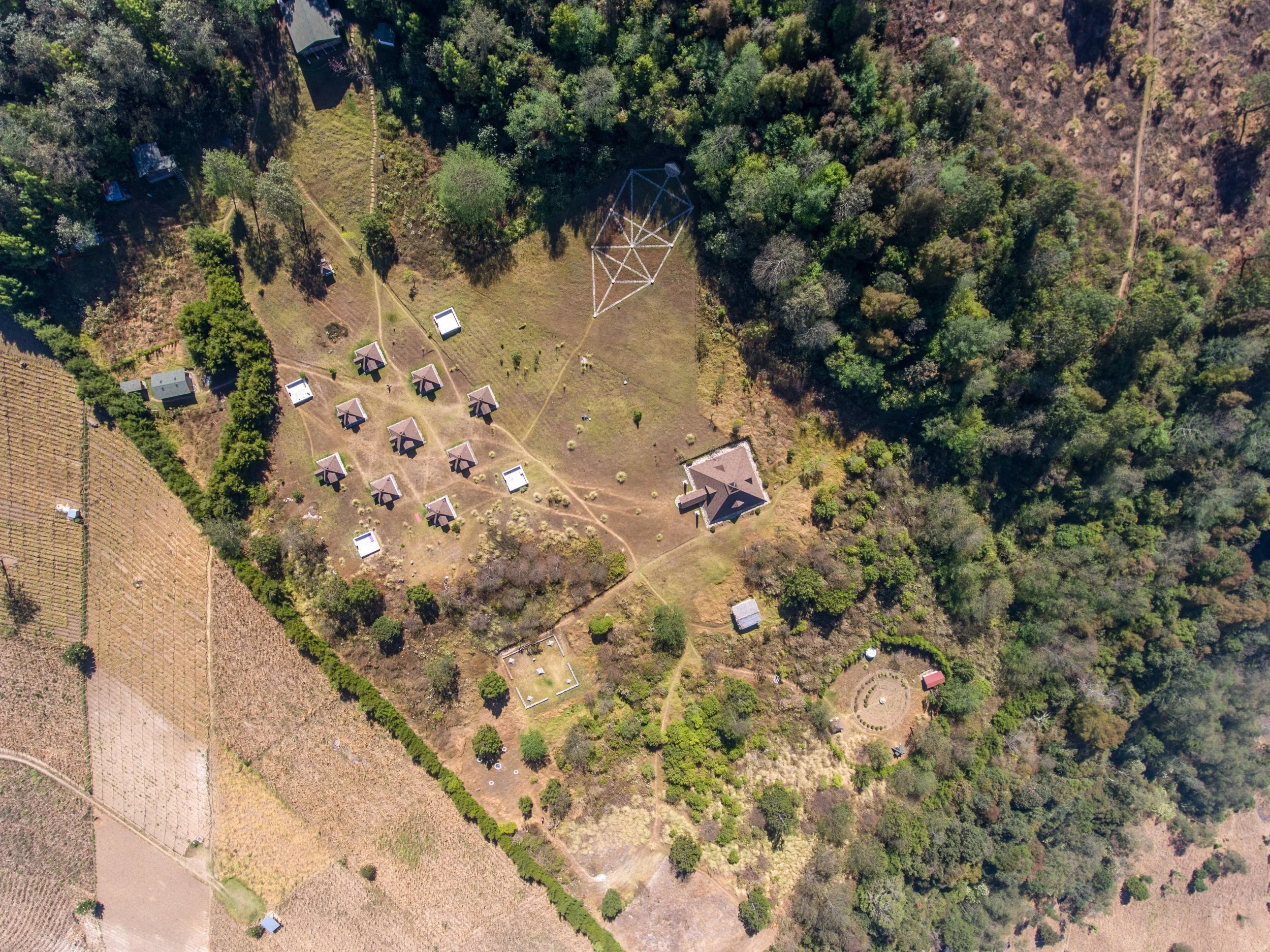 Vista aérea de un área verde con árboles, caminos, una estructura de cuerda, varias cabañas y cultivos agrícolas en los alrededores.