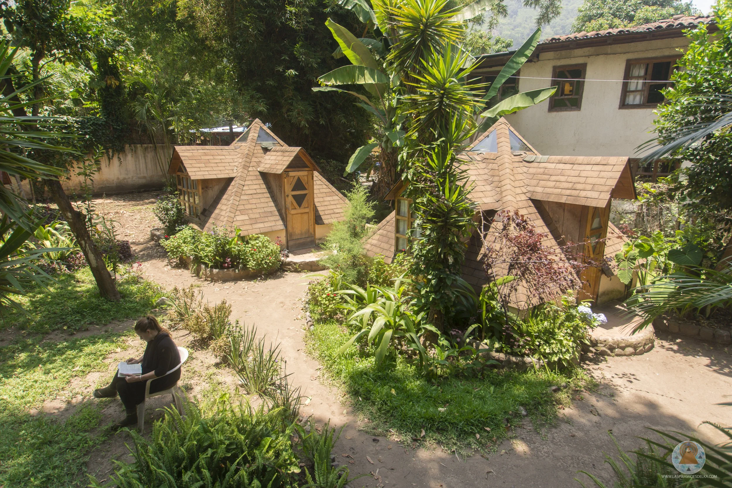 Jardín con plantas tropicales y dos pequeñas casas de madera con techos inclinados y ventanas en medio de vegetación densa. En primer plano, una persona sentada en una silla leyendo un libro.