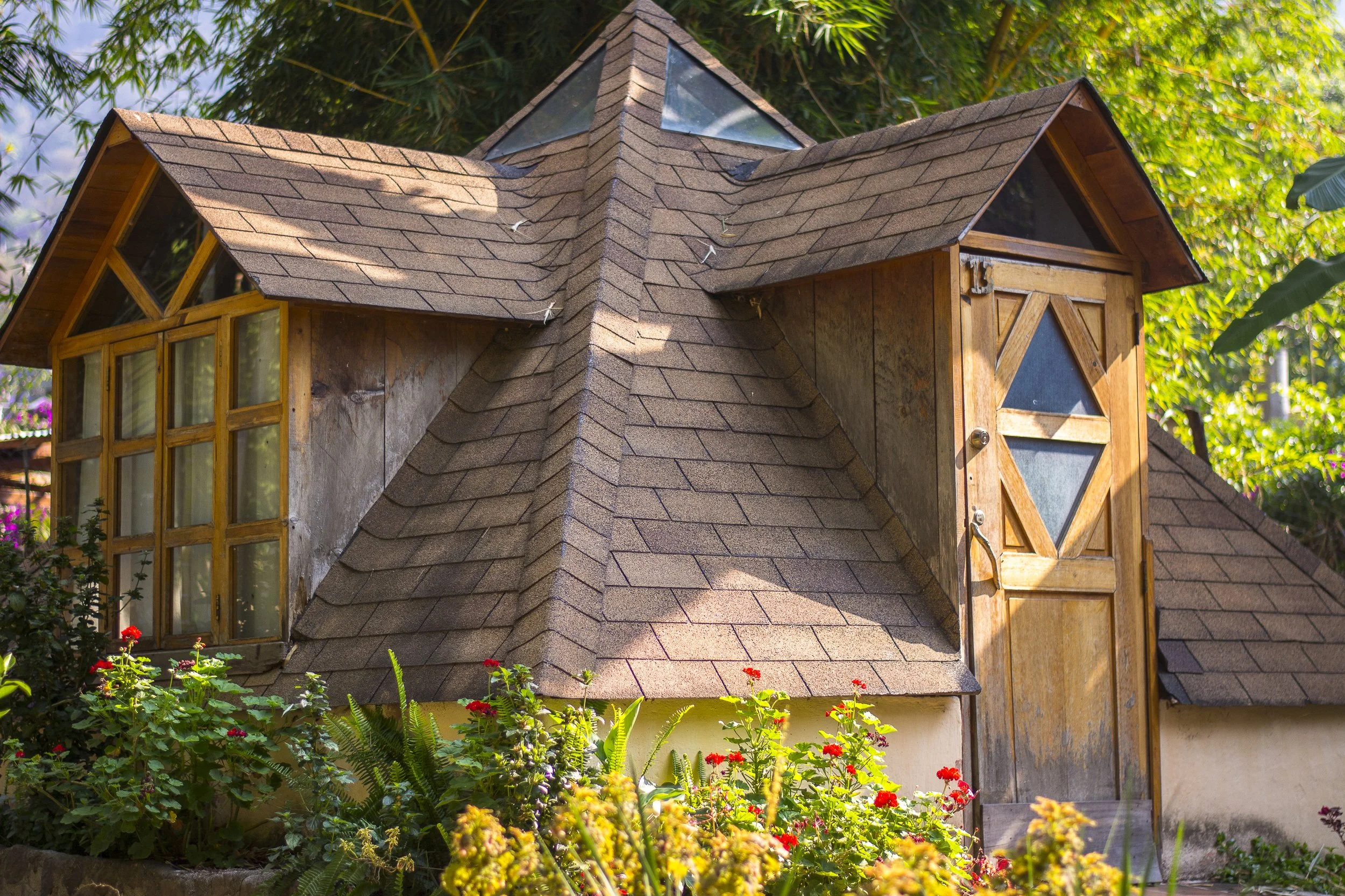 Casa de madera con tejado de tejas y ventanas en forma de triángulo, rodeada de flores y plantas verdes, con árboles en el fondo.