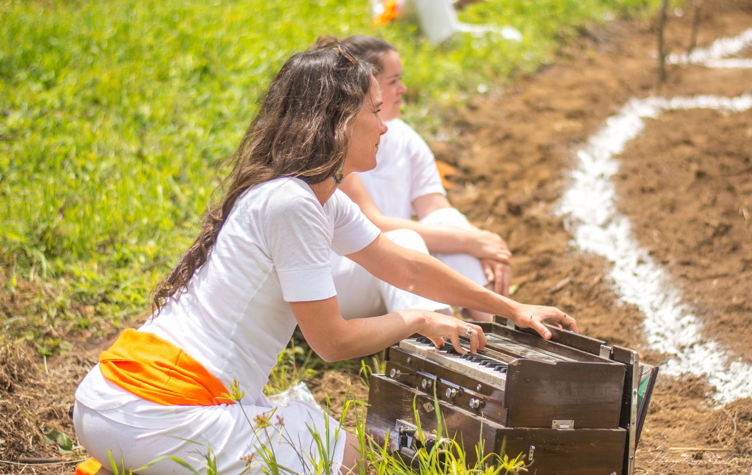 Mujeres participando en una ceremonia espiritual al aire libre, tocando un piano y sentadas en la tierra en un campo con vegetación.