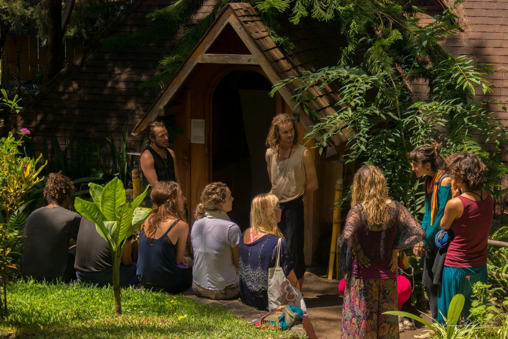 Personas participando en una ceremonia o charla al aire libre frente a una pequeña estructura de madera en un entorno natural con abundante vegetación.