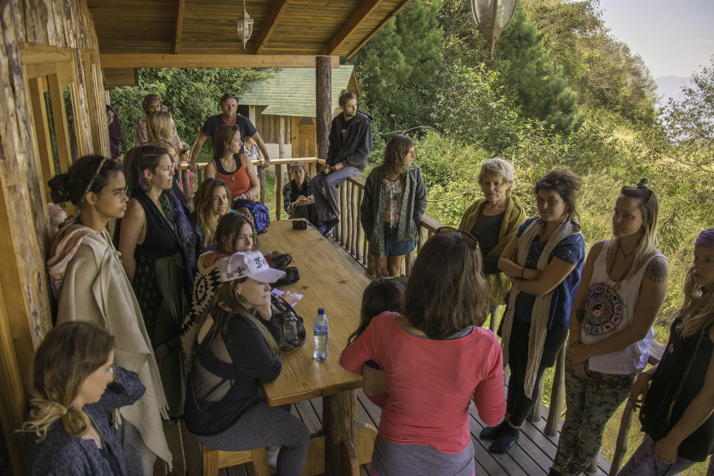 Grupo de personas en una terraza de madera rodeada de árboles, participando en una reunión o charla al aire libre, con algunos tomando agua y otros conversando.