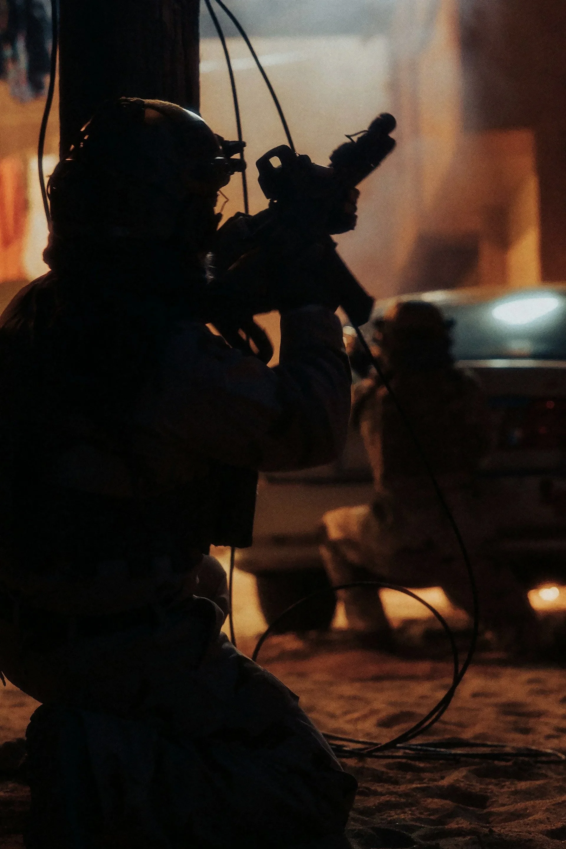 Silhouette of a person working with wires in a dimly lit environment, possibly on a construction site or in a workshop, with a blurred background that includes a vehicle and some structures.