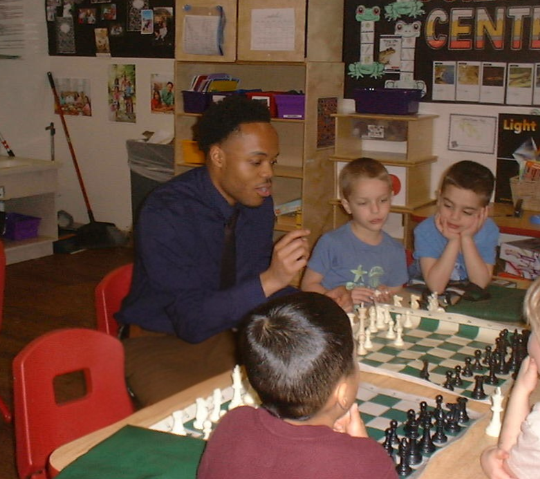 A man teaching a chess game to young children in a classroom setting with educational posters and shelves in the background.