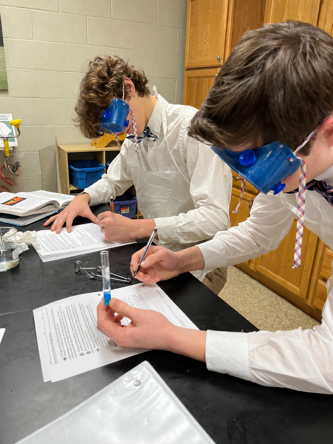 Students in safety goggles conducting a science experiment in a classroom, wearing lab coats, and working with papers, test tubes, and scientific equipment on the table.