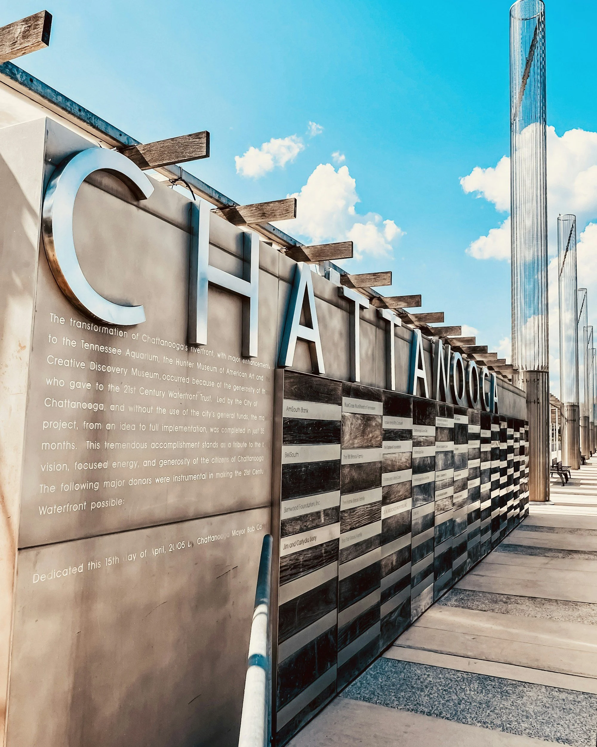 Part of a building with large silver letters spelling out 'CHATTANOOGA'. There is a wall with informational text and images, and tall, reflective sculptures against a bright blue sky with some clouds.