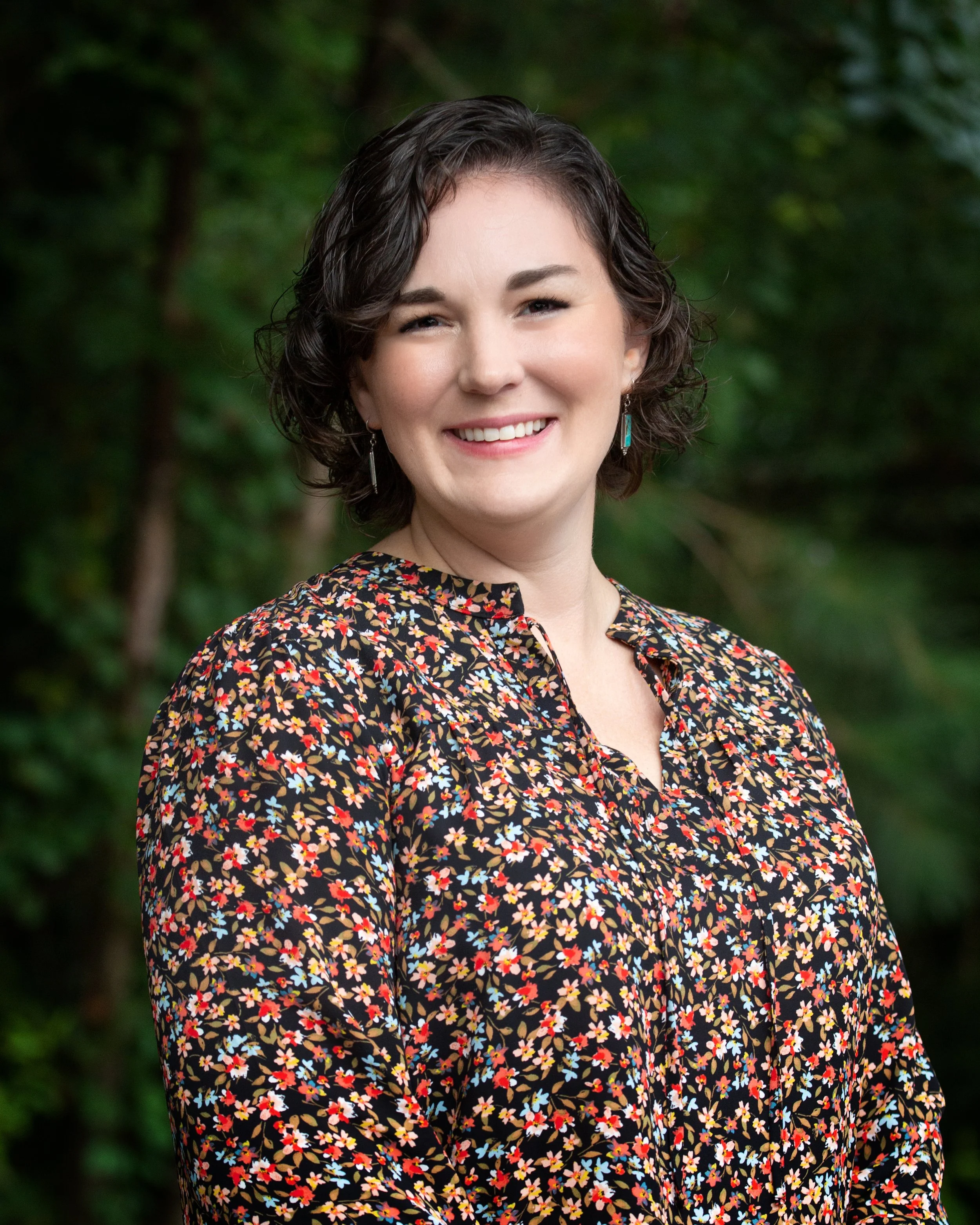 A woman with short, dark, curly hair smiling outdoors with a blurred green background, wearing a black dress with a multicolored floral pattern and turquoise earrings.
