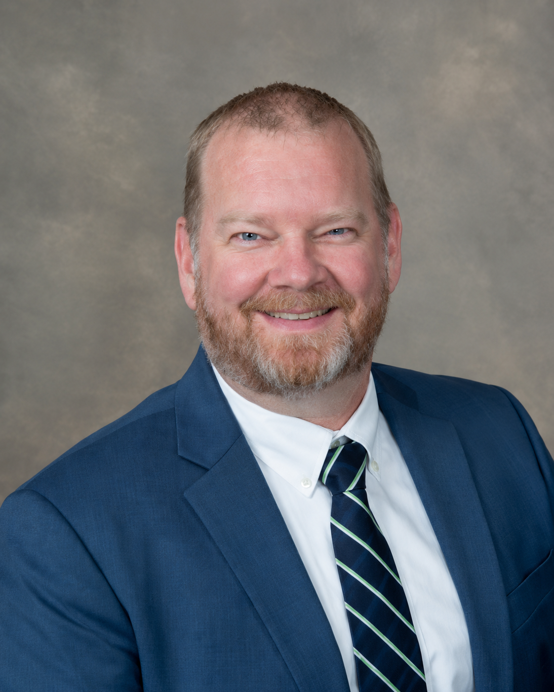 Professional headshot of a man with light skin, short light brown hair, a beard, wearing a navy suit, white shirt, and a navy and green striped tie, smiling against a neutral background.