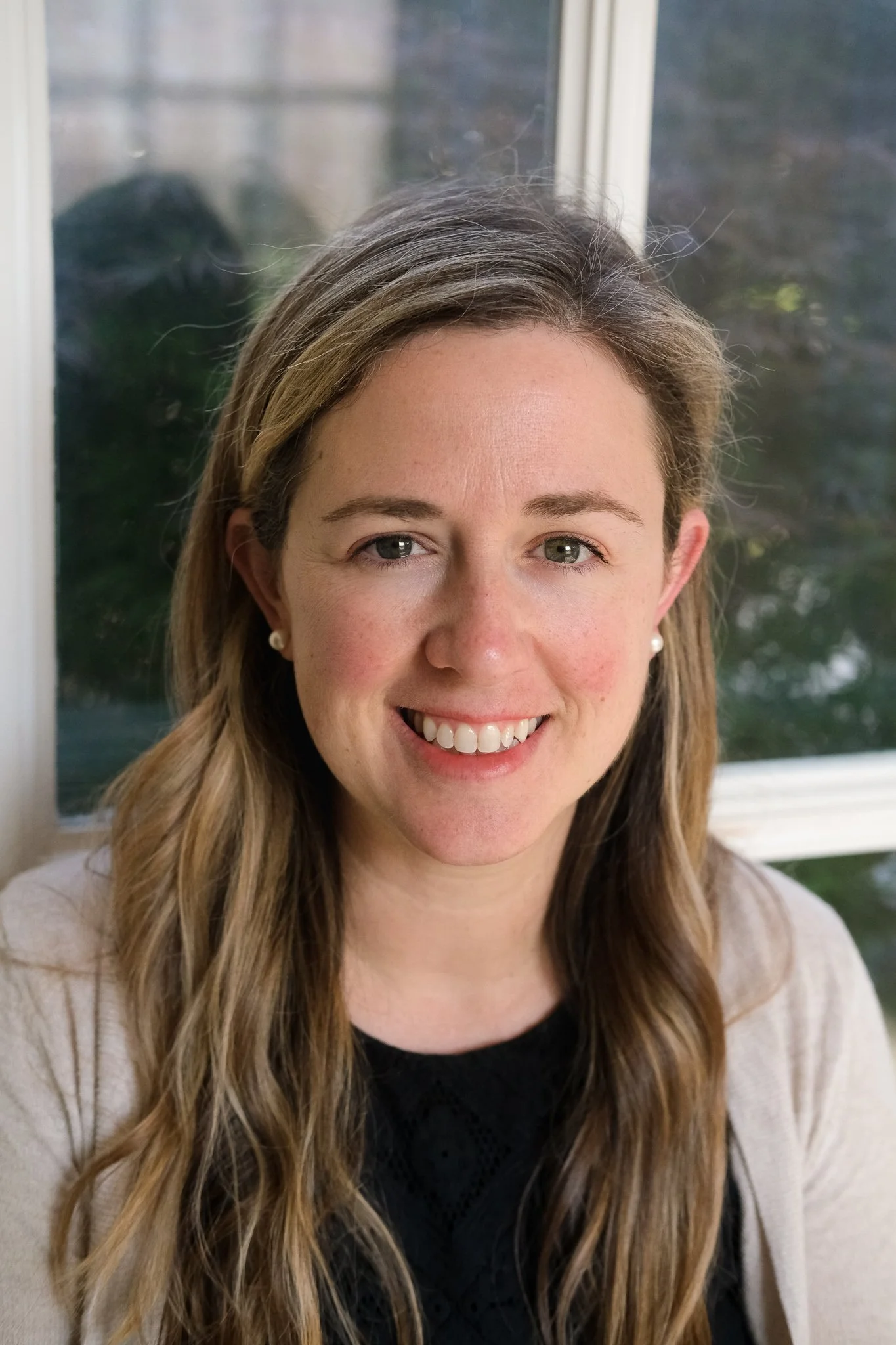 A woman smiling while sitting near a window with a blurry outdoor view.