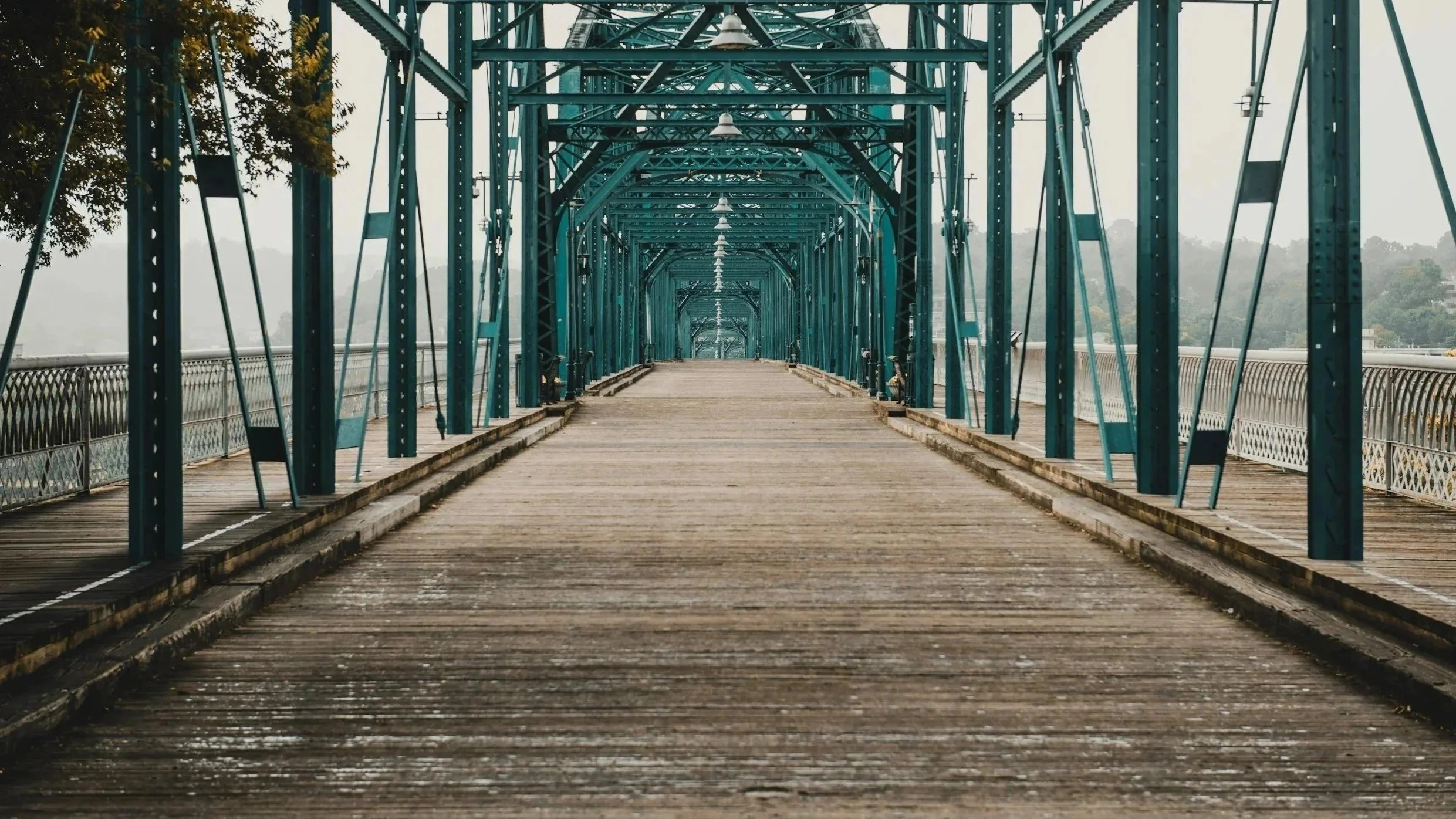 The image depicts a foggy, blue-green metal bridge or walkway with a wooden floor. It is Chattanooga's walking bridge over the Tennessee River.