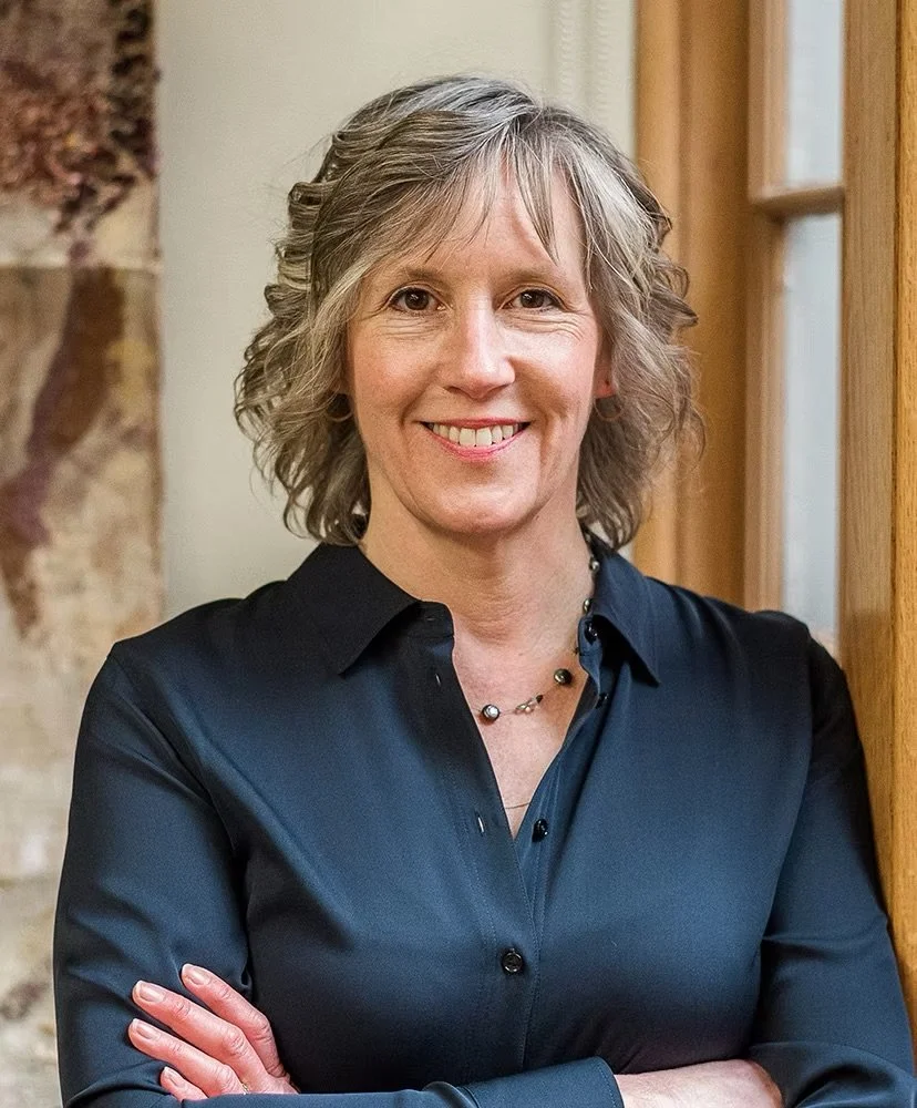 A woman with curly gray hair smiling, wearing a black shirt and a necklace, standing indoors near a wooden door and stone wall.