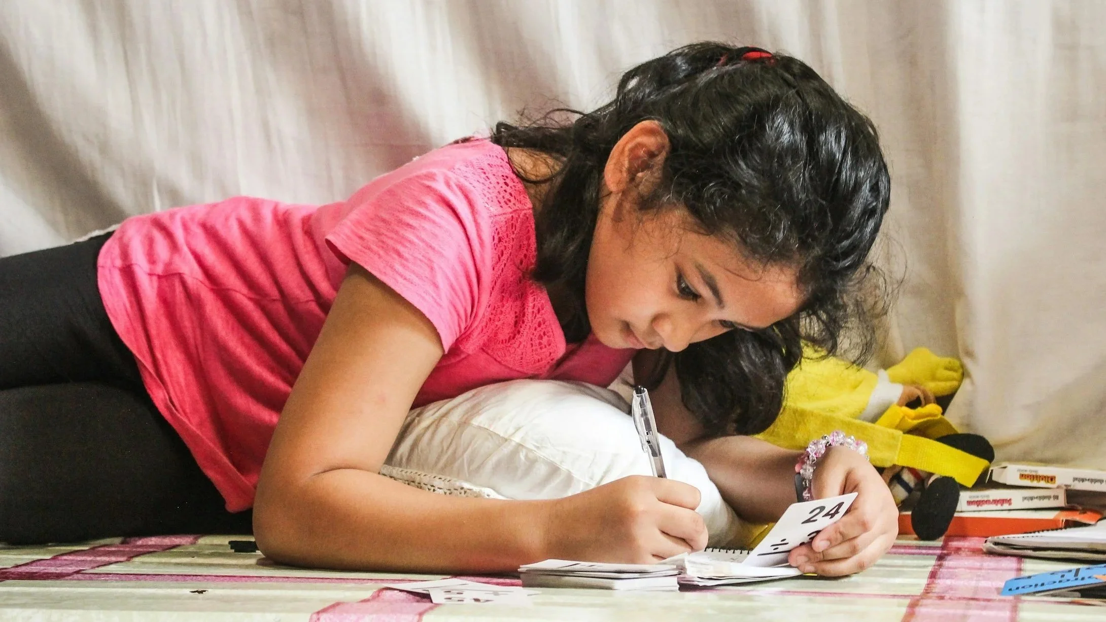 A woman with curly black hair and wearing a pink shirt is lying on the floor, studying or working with papers, a pen, and a small sign with the number 24.