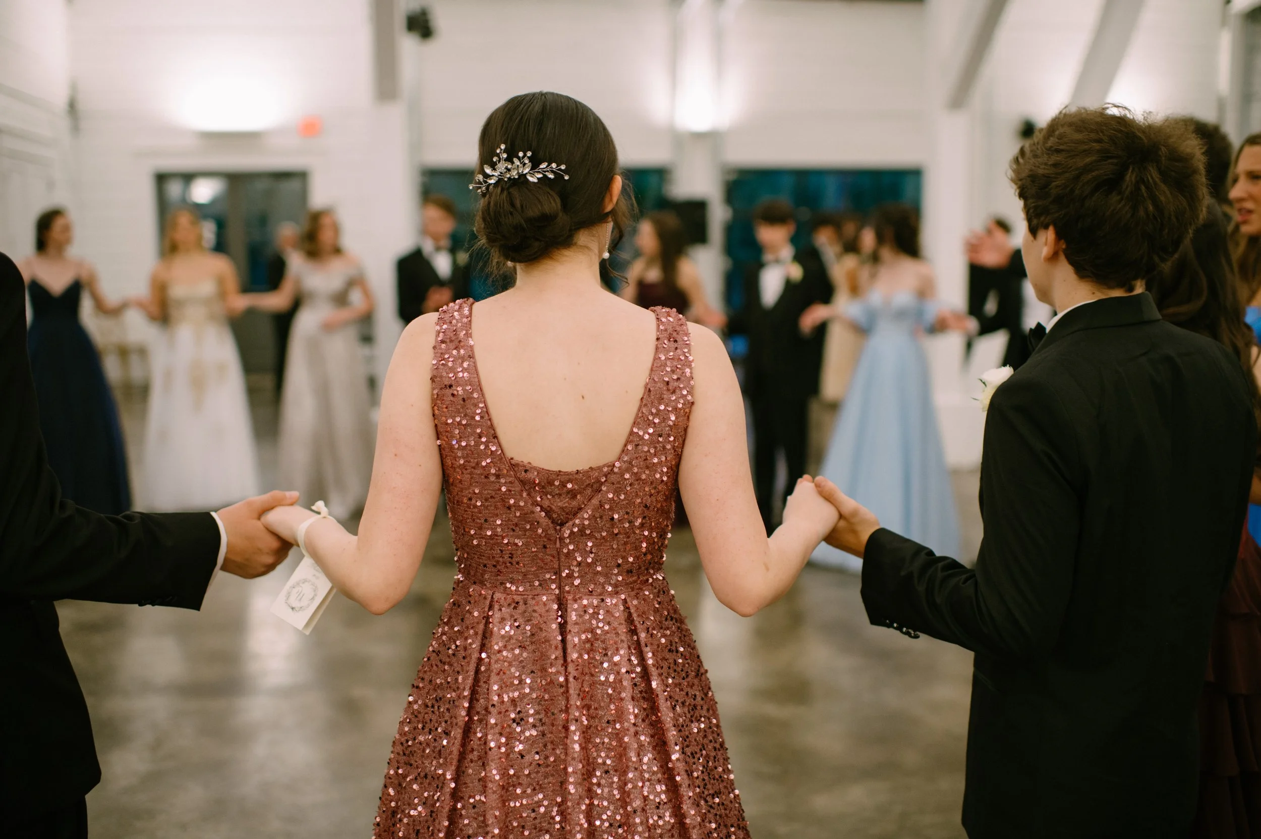 A woman in a pink, sequined gown dancing with a man and woman during a formal event or wedding reception, holding hands in a circle with other guests.
