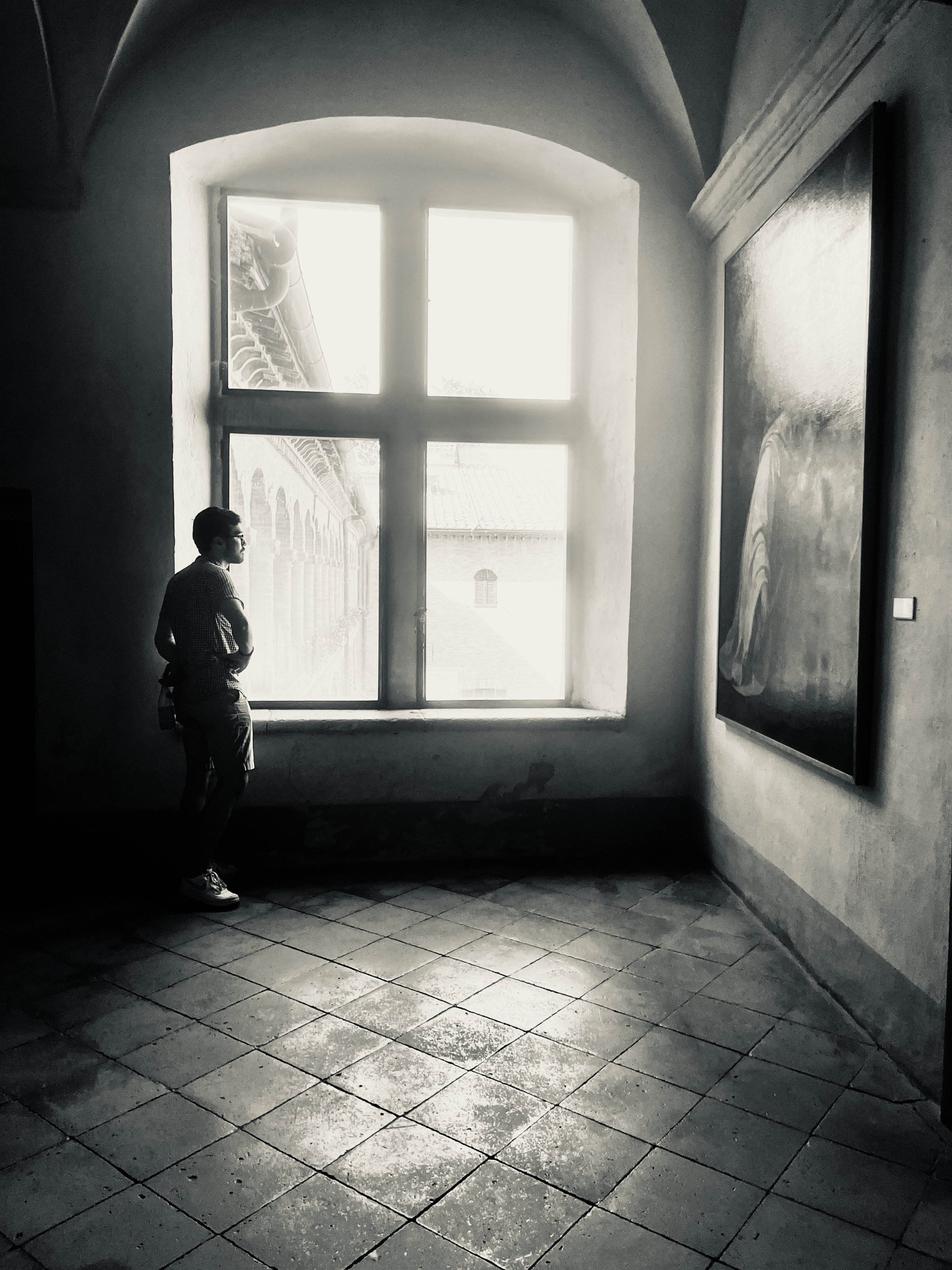 A person standing inside a building near a large window, with sunlight streaming in, casting shadows on the tiled floor and a wall with a large painting.