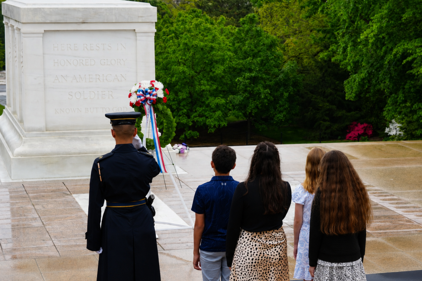 People standing at attention in front of a war memorial with a soldier laying a wreath during a memorial ceremony, surrounded by trees and flowers.