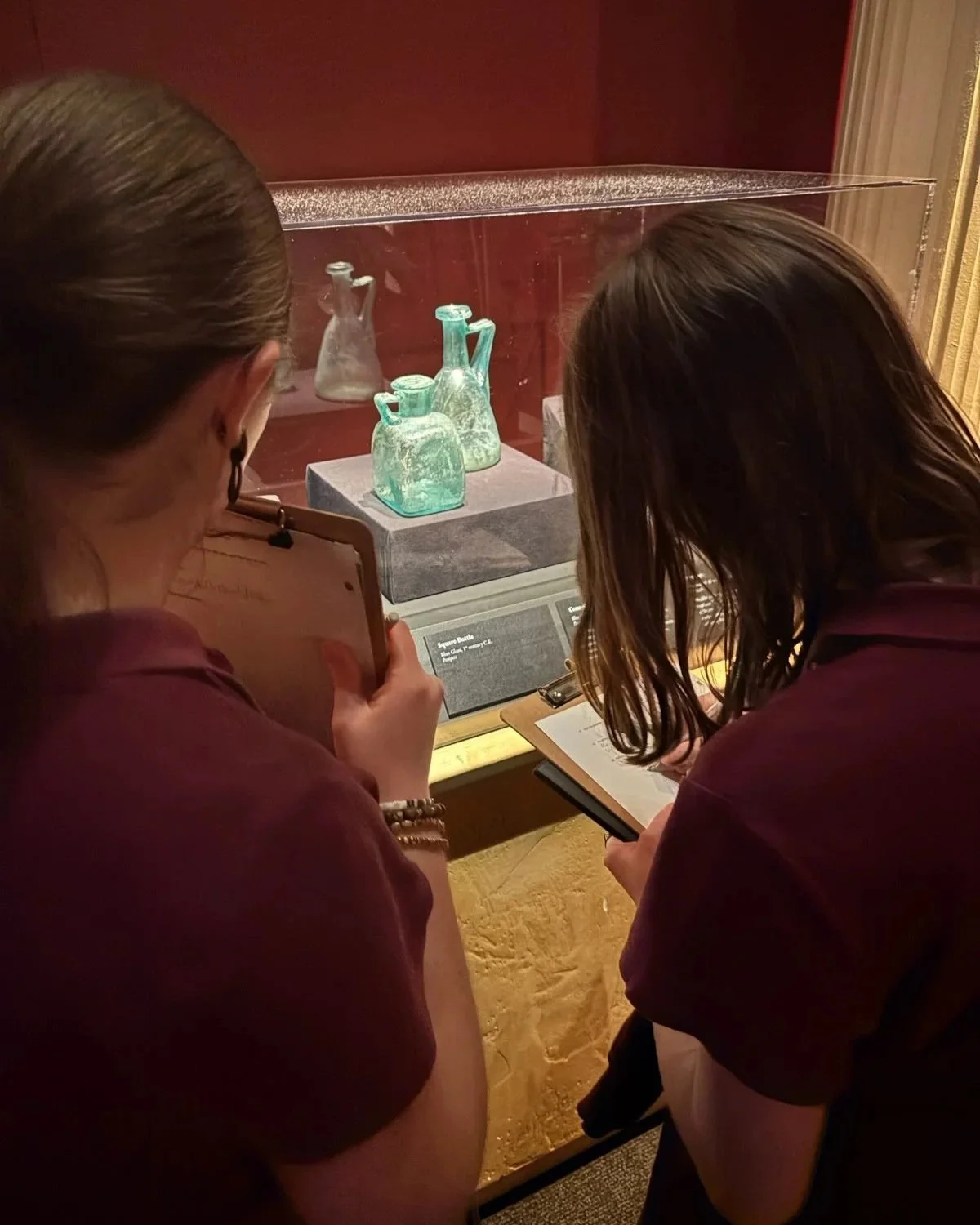 Two women viewing ancient glass bottles in a museum display case, taking notes and discussing the exhibit.