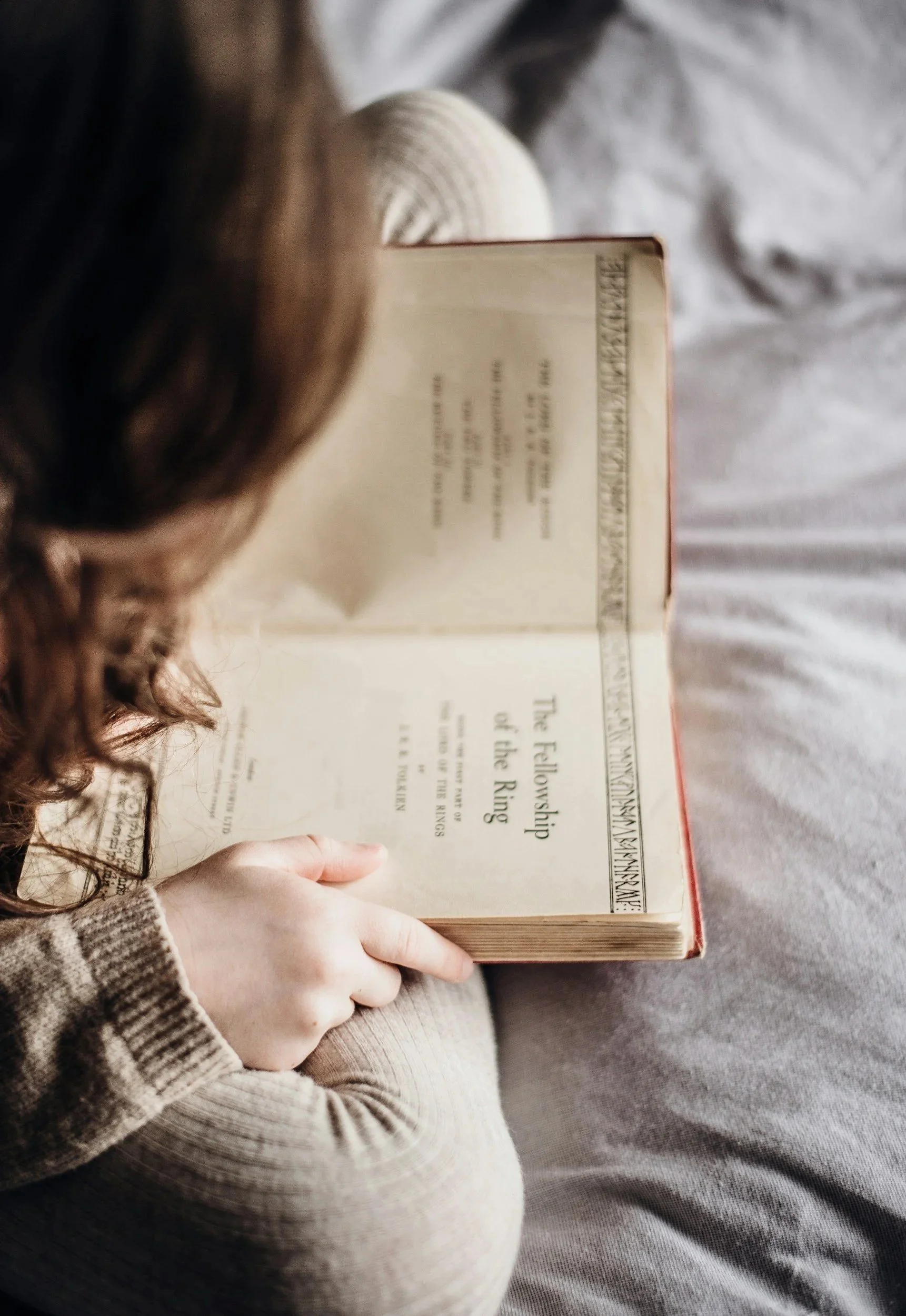 A person with curly hair and a striped sweater holding an open book titled 'The Fellowship of the Ring' by J.R.R. Tolkien, sitting on a soft surface.