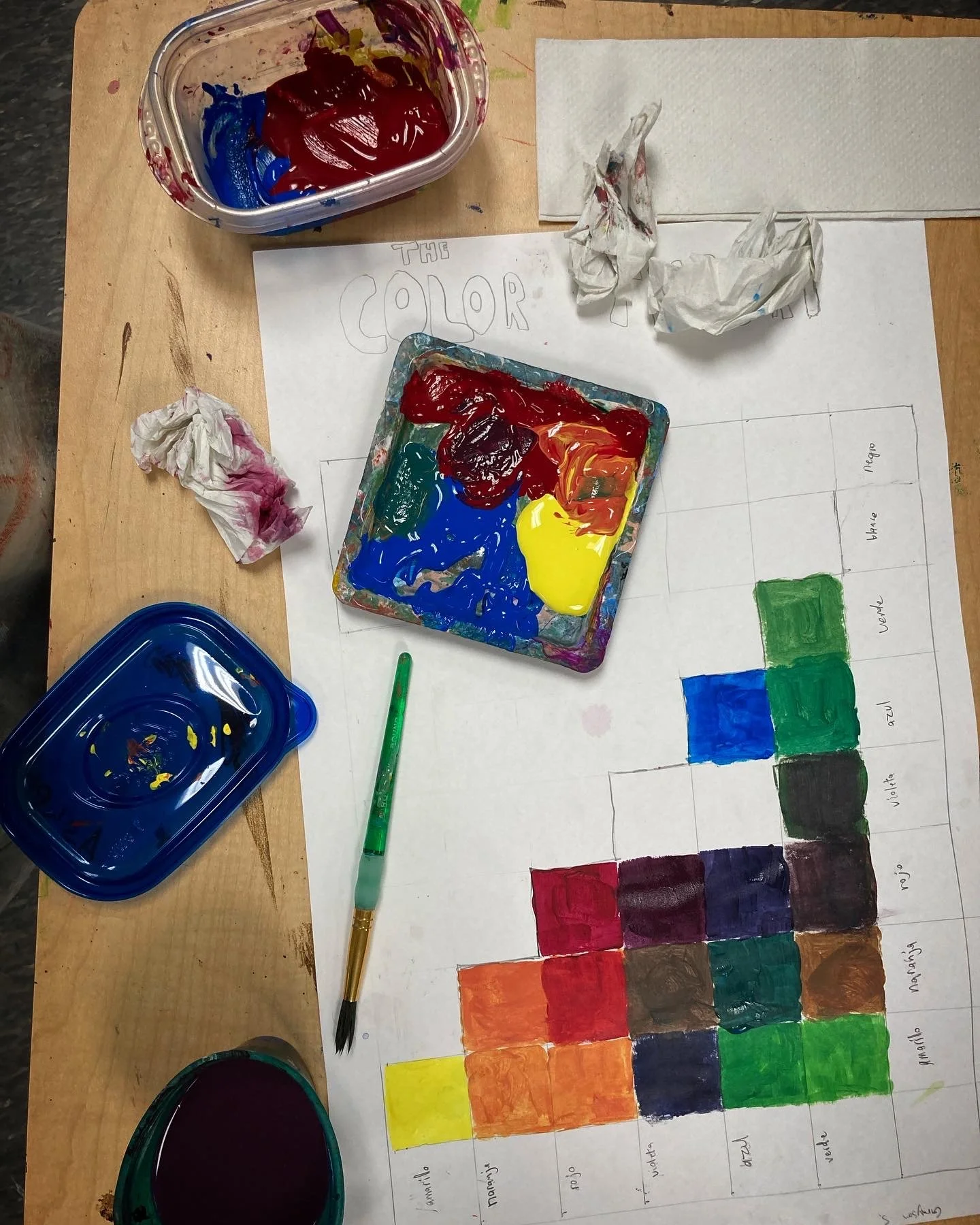 A workspace with a color chart, paint supplies, and a sheet of paper with the word 'COLOR' and a color palette. There are multiple crumpled paper towels stained with paint, and containers of mixed paint. A green paintbrush rests on the paper.