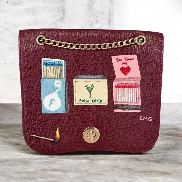 A red handbag decorated with embroidered patches of a matchbox with a fuse, a martini glass with the words "Extra Dirty," a pink matchbook with a heart and the words "You Know my," and a small yellow flame at the bottom left. The bag has a gold chain handle and a turn-lock closure.