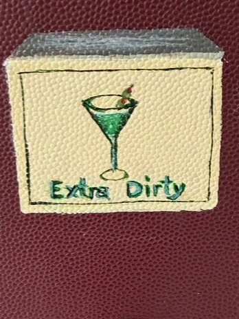 Embroidery of a green cocktail glass with a cherry and lime wedge, with the phrase 'Extra Dirty' underneath, on a red textured fabric background.
