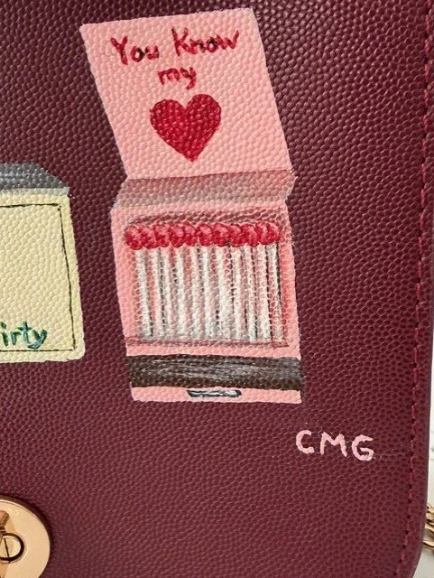 Tiny pink matchbox with a red heart and the words 'You know my' above it, sitting on a maroon surface with the initials 'CMG' nearby.