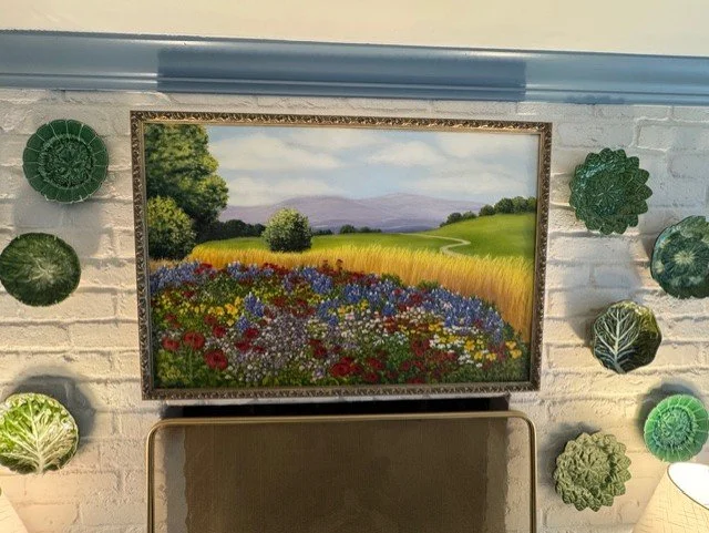 A framed landscape painting of a colorful flower field with trees and mountains on a white brick wall, surrounded by green decorative plates and a partial view of a beige chair and lamp.