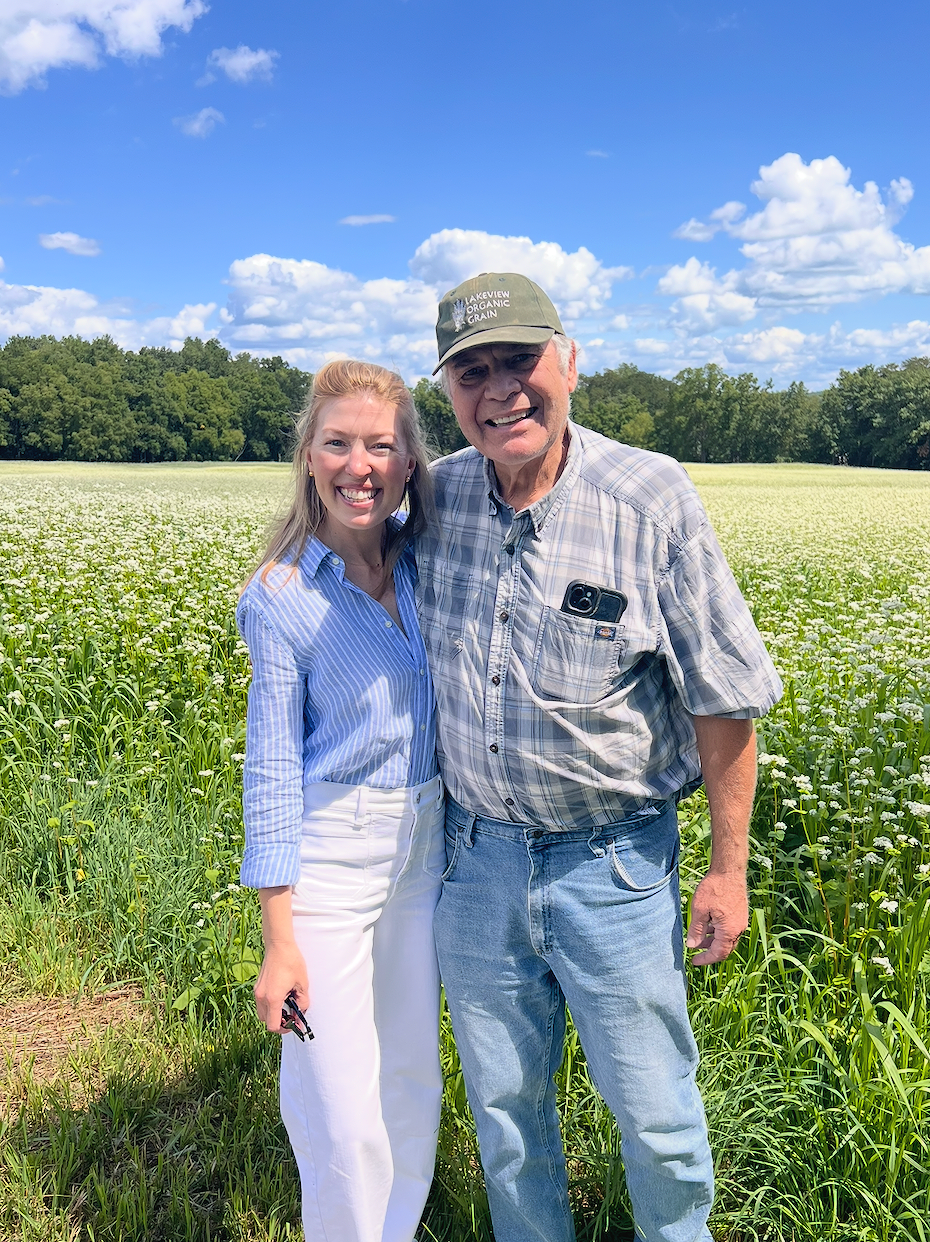 A woman and an older man standing together in a field of white flowers under a blue sky with scattered clouds, smiling at the camera.