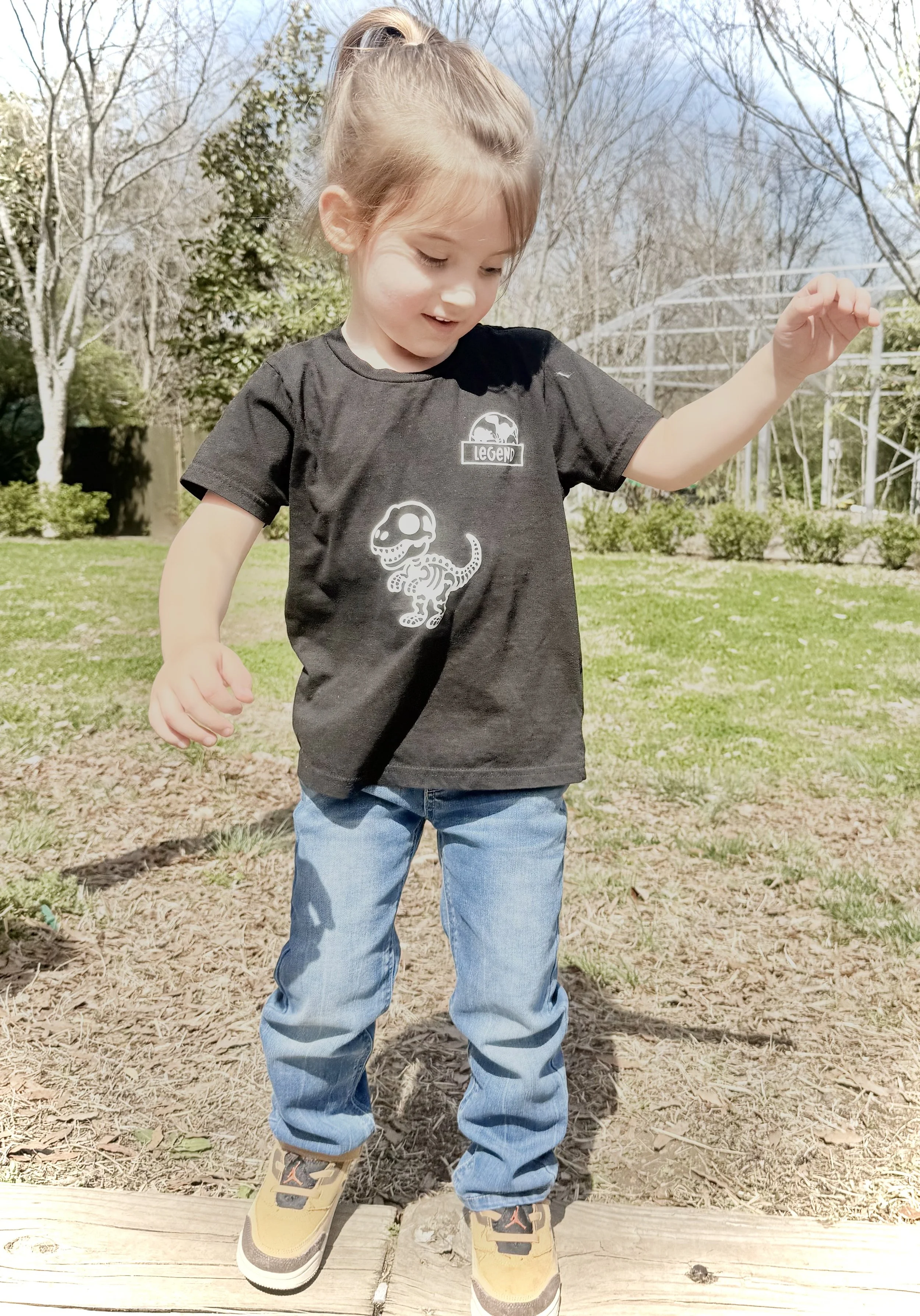 A young boy standing outdoors on a sunny day, wearing a black T-shirt with a dinosaur graphic and sneakers, with trees and a metal structure in the background.