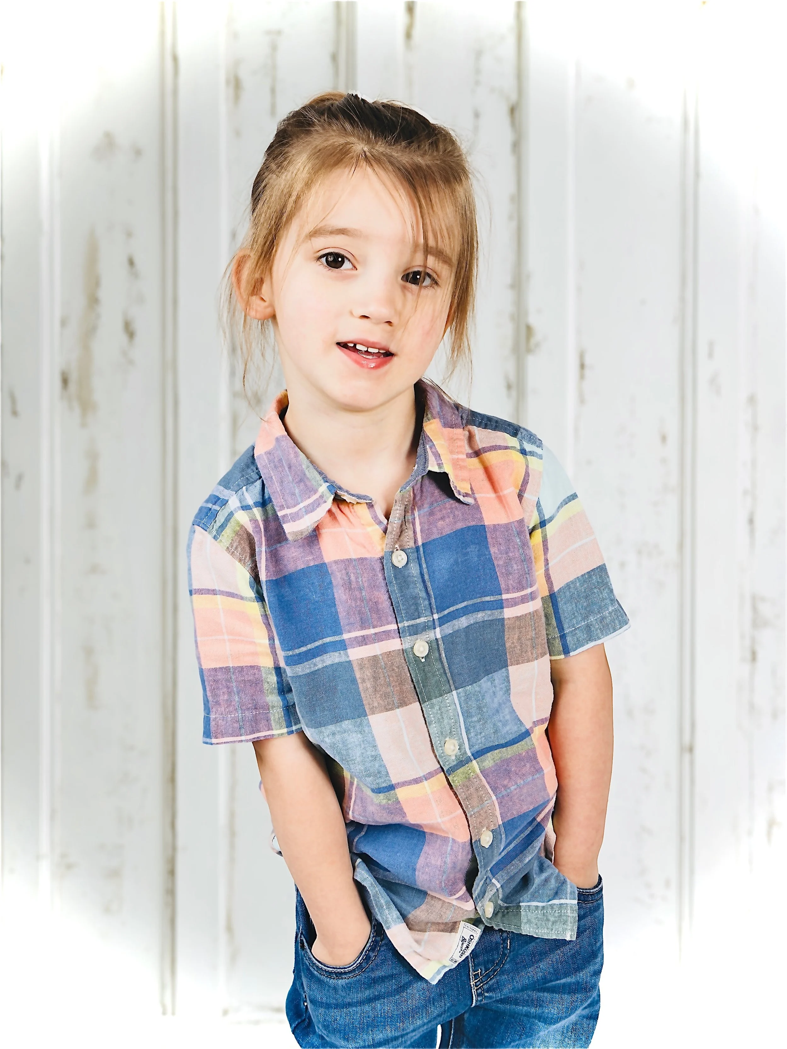 A young girl with brown hair tied back, wearing a multi-colored plaid shirt with short sleeves and blue jeans, standing in front of a white wooden wall.