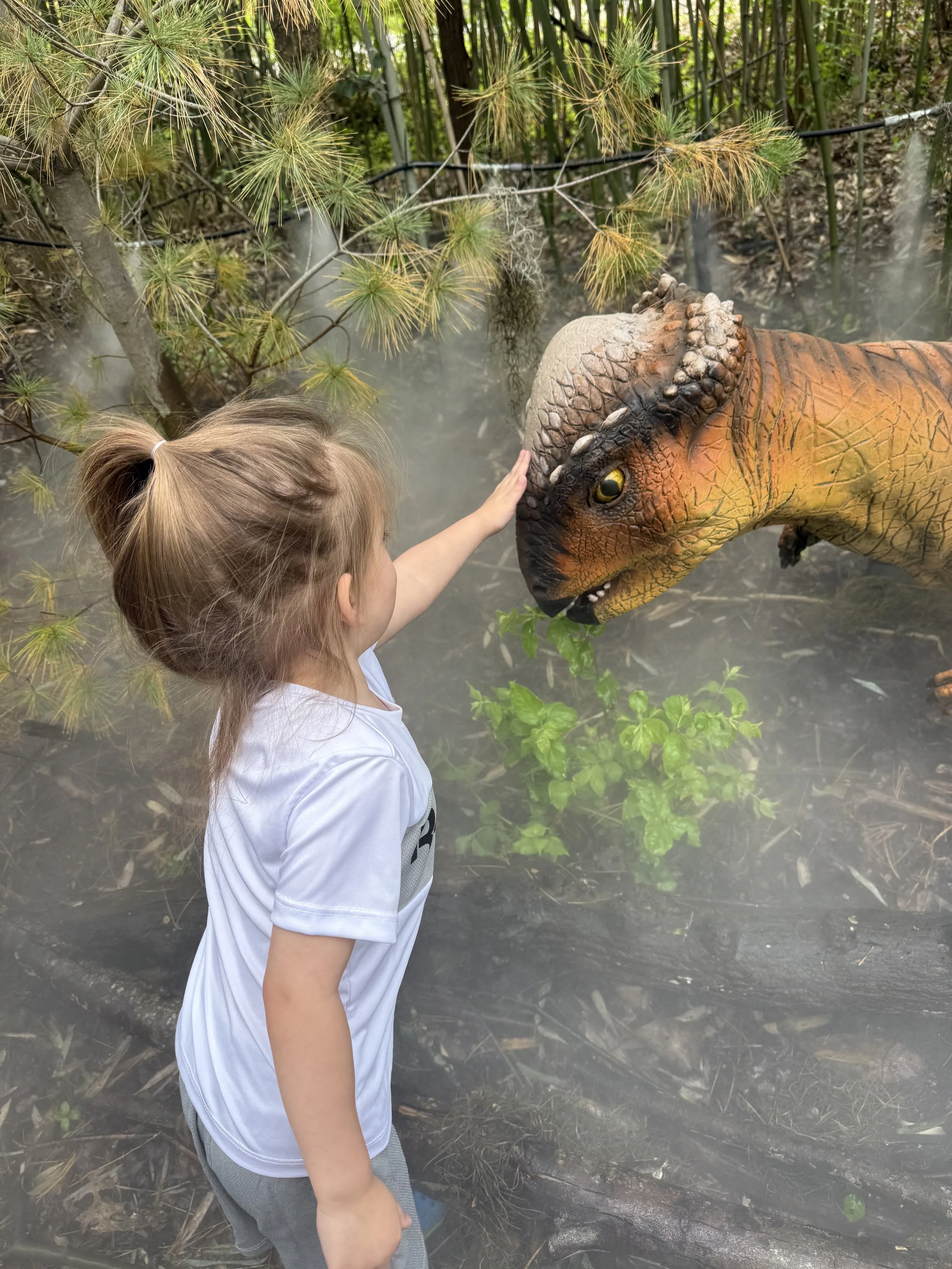 A young girl petting a lifelike dinosaur model in a forest setting.