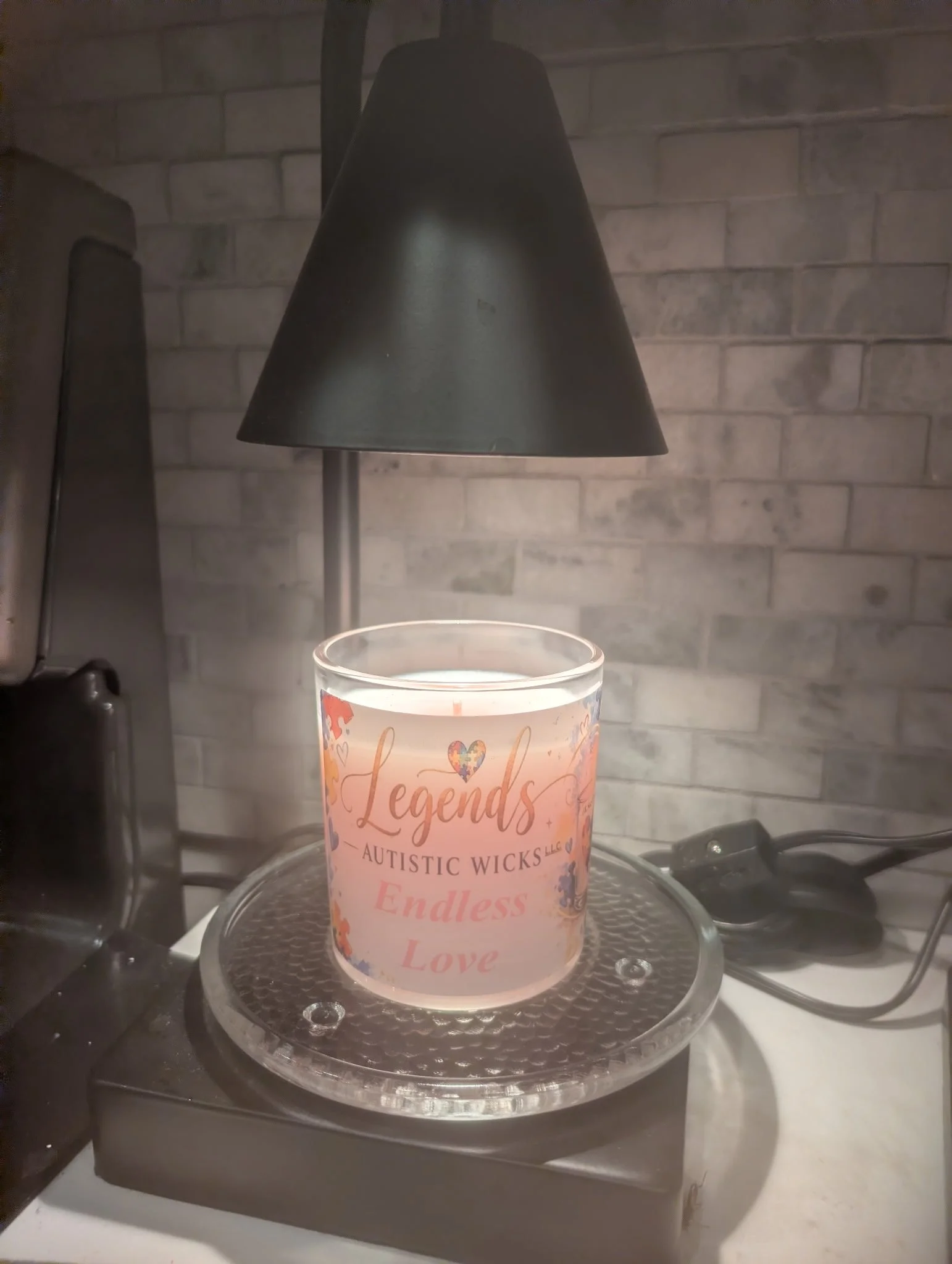 A lit candle with a pink label featuring words like 'Legends,' 'Autistic Wicks,' and 'Endless Love,' placed on a clear glass holder underneath a black lampshade.