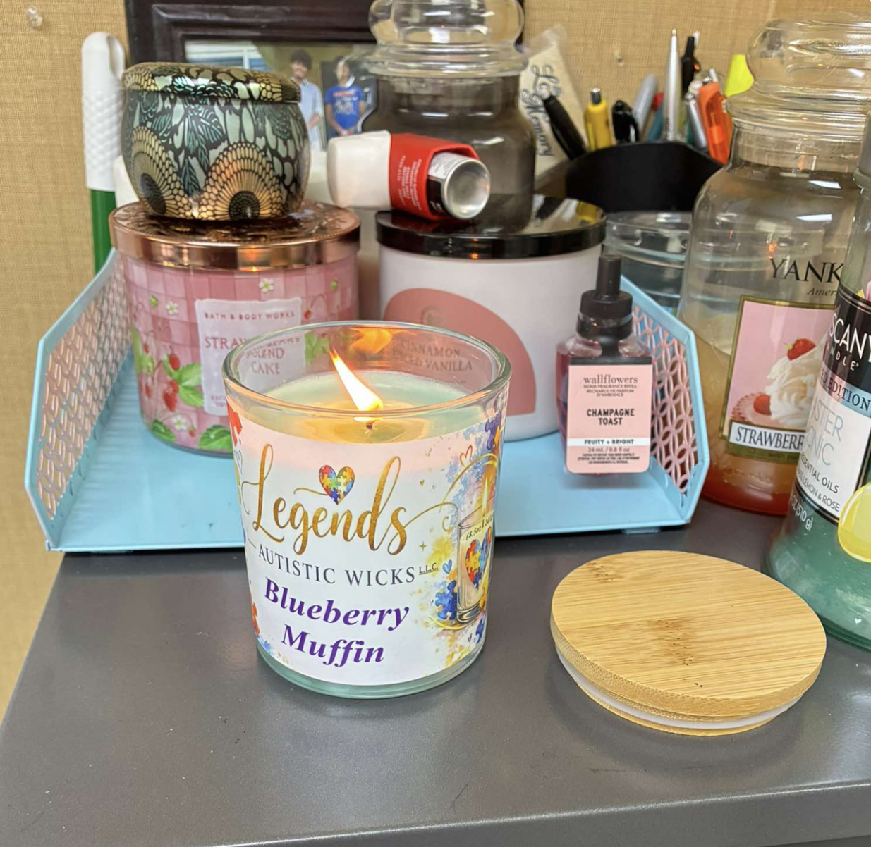 A lit scented candle labeled 'Legends Autistic Wicks Blueberry Muffin' sits on a table with various candles, jars, and containers in the background.