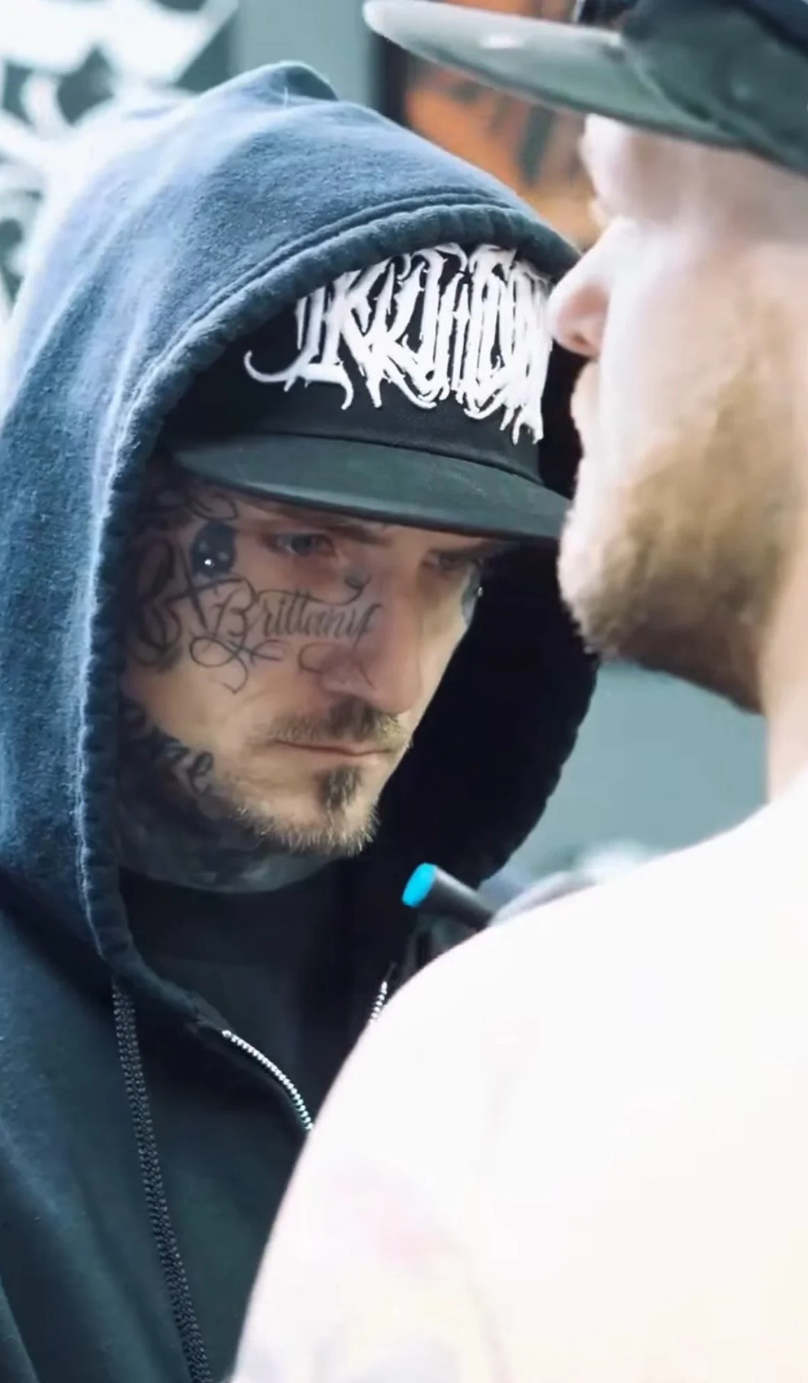 A close-up of a man with tattoos, wearing a hoodie and a hat, looking intently at another person.