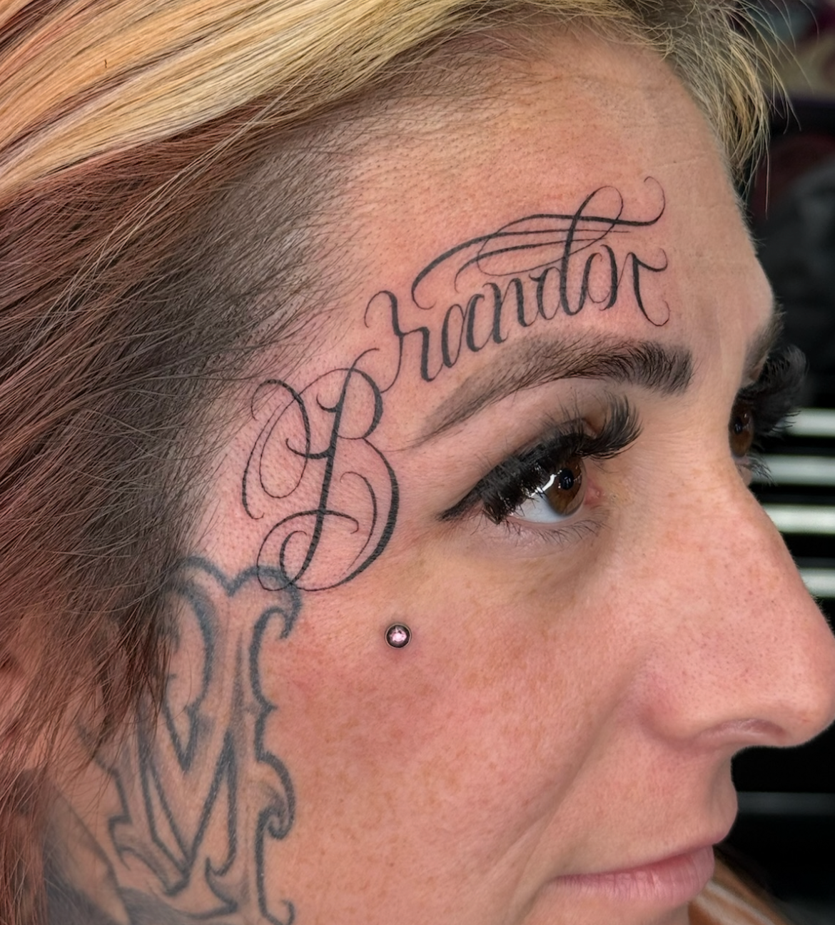 Close-up of a woman's face, showing tattoos including the word 'Brandon' above her right eyebrow, a decorative tattoo on her cheek, and another tattoo near her hairline. She has long, dark eyelashes and a small facial piercing.