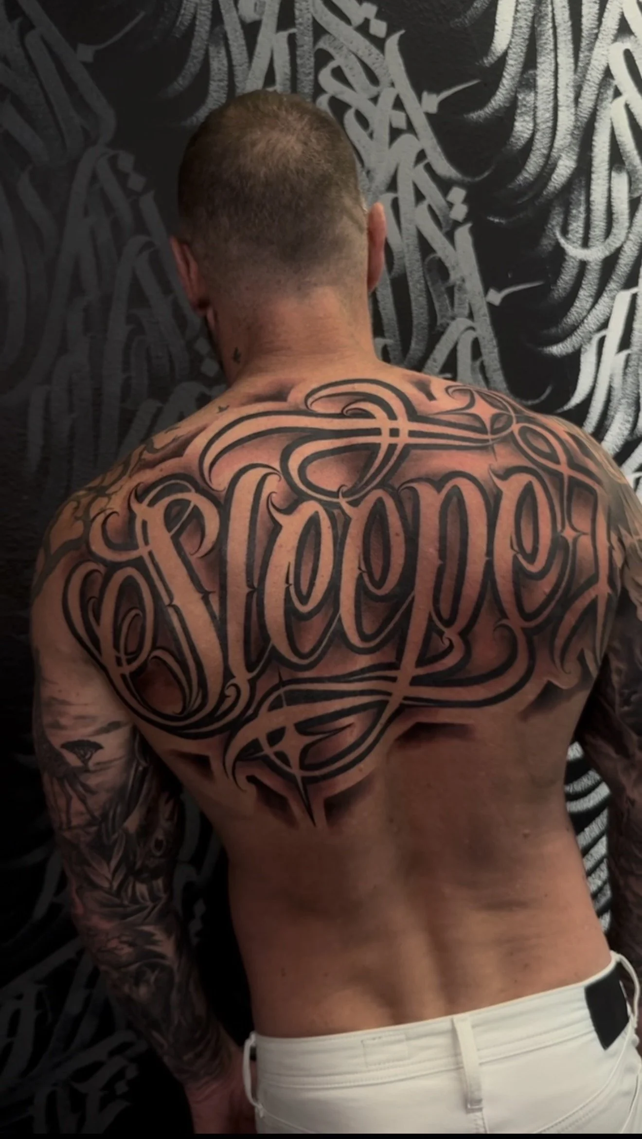 Back view of a man with tattoos, featuring a large black and gray tattoo of the word 'respect' in cursive script on his chest, lying face down on a surface with a black and white abstract background.