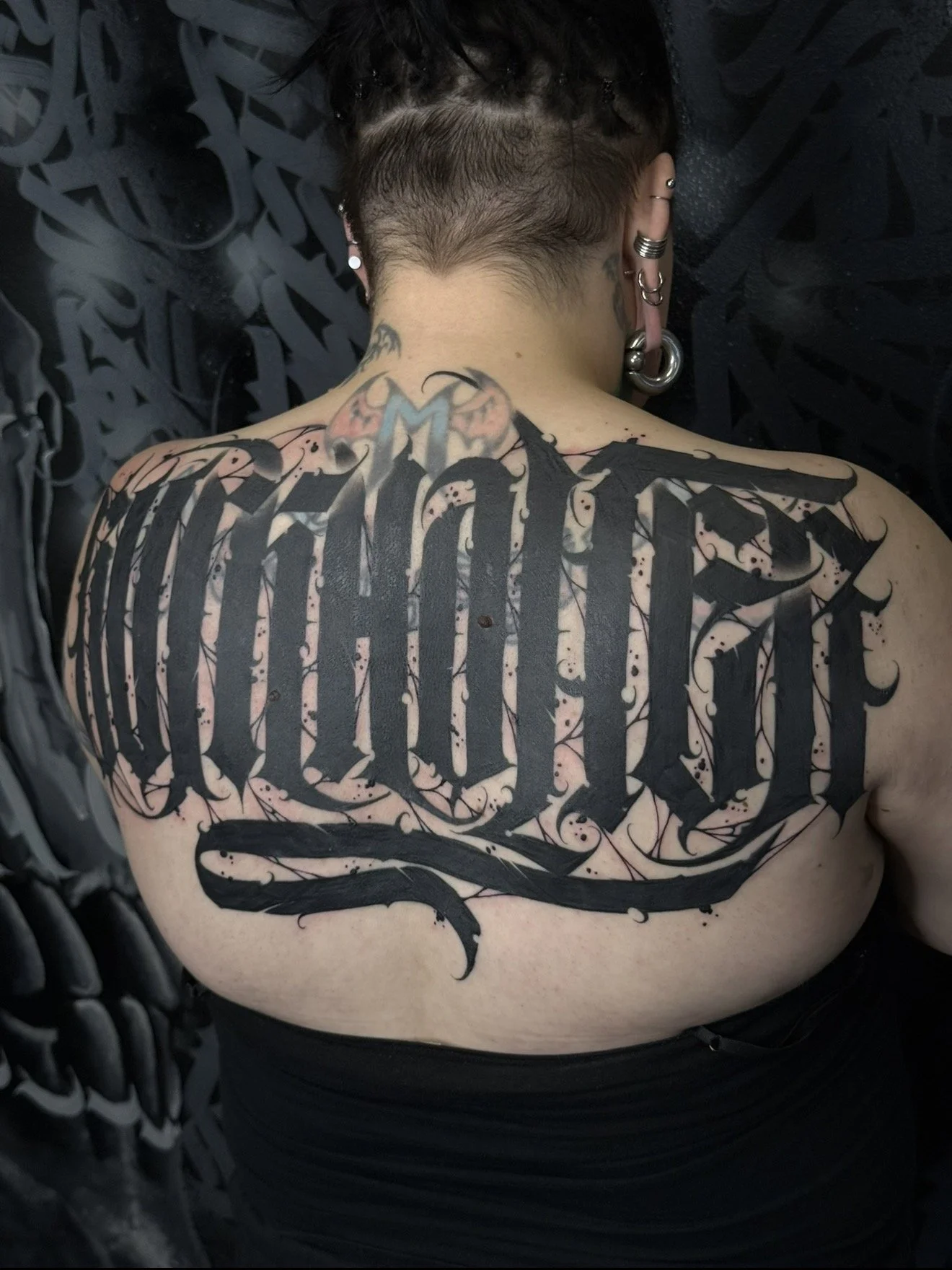 Back of a person with a large black tattoo in gothic style text across their back and smaller tatted designs on the neck and ears.
