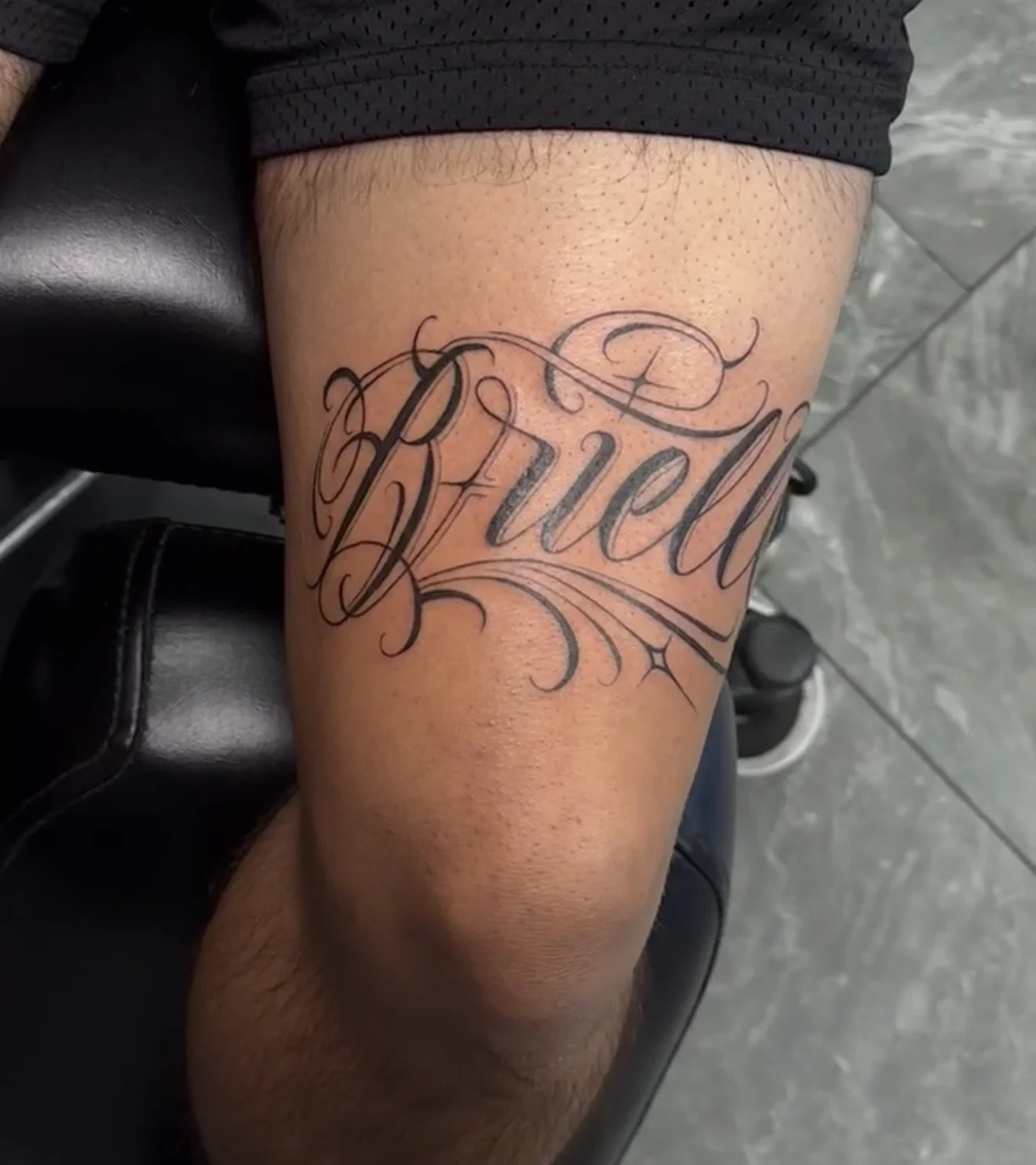 Tattoo of the word 'Drew' in elaborate cursive script on a person's thigh.