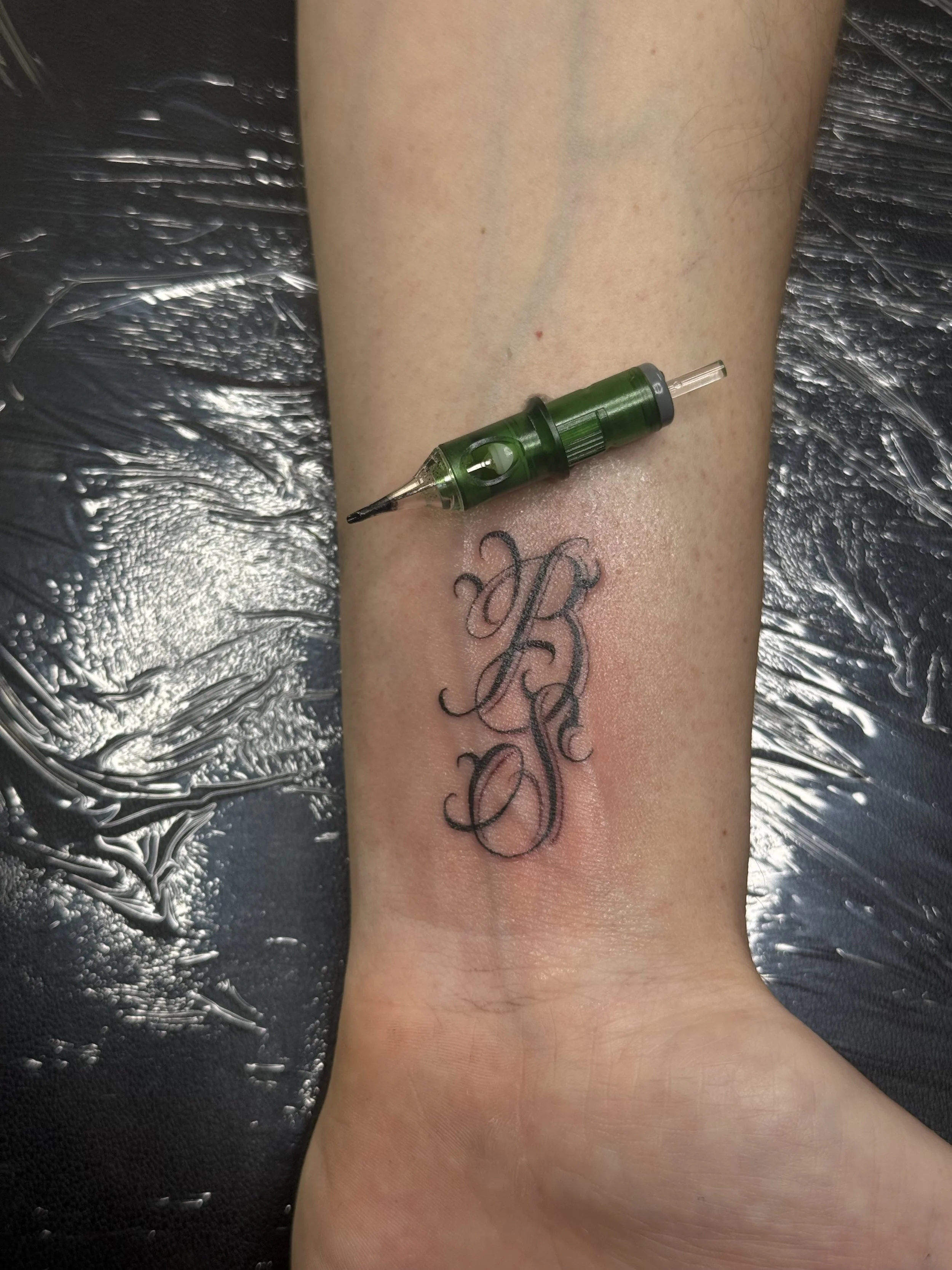 A fresh tattoo of ornate intertwined initials 'K' and 'R' is being inked on a person's ankle, with a tattoo machine resting on the skin.